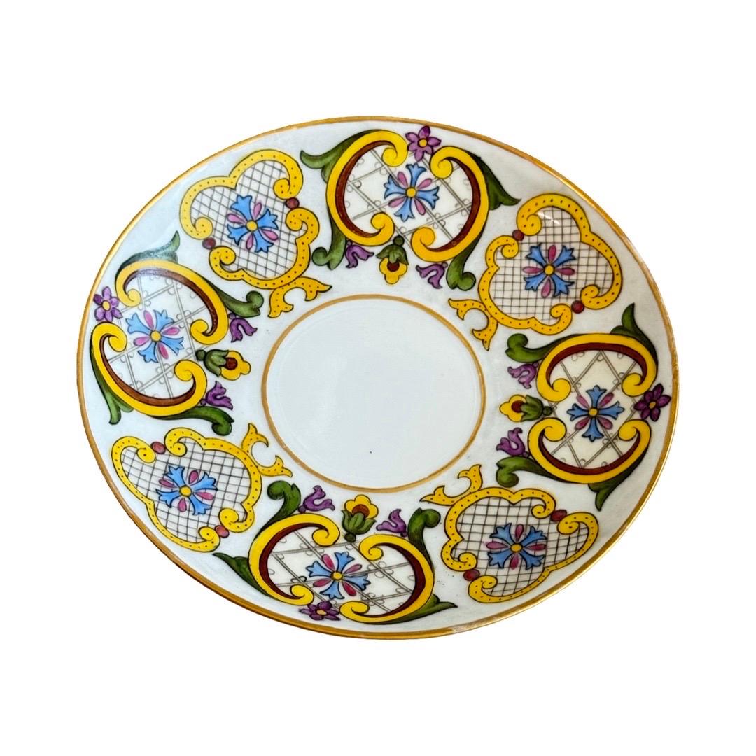A refined antique porcelain cup and saucer set, richly hand-painted in a vibrant palette of blue, yellow, pink, and green. The intricate design features floral motifs, ornamental grids, and gold accents throughout. The handle is delicately shaped