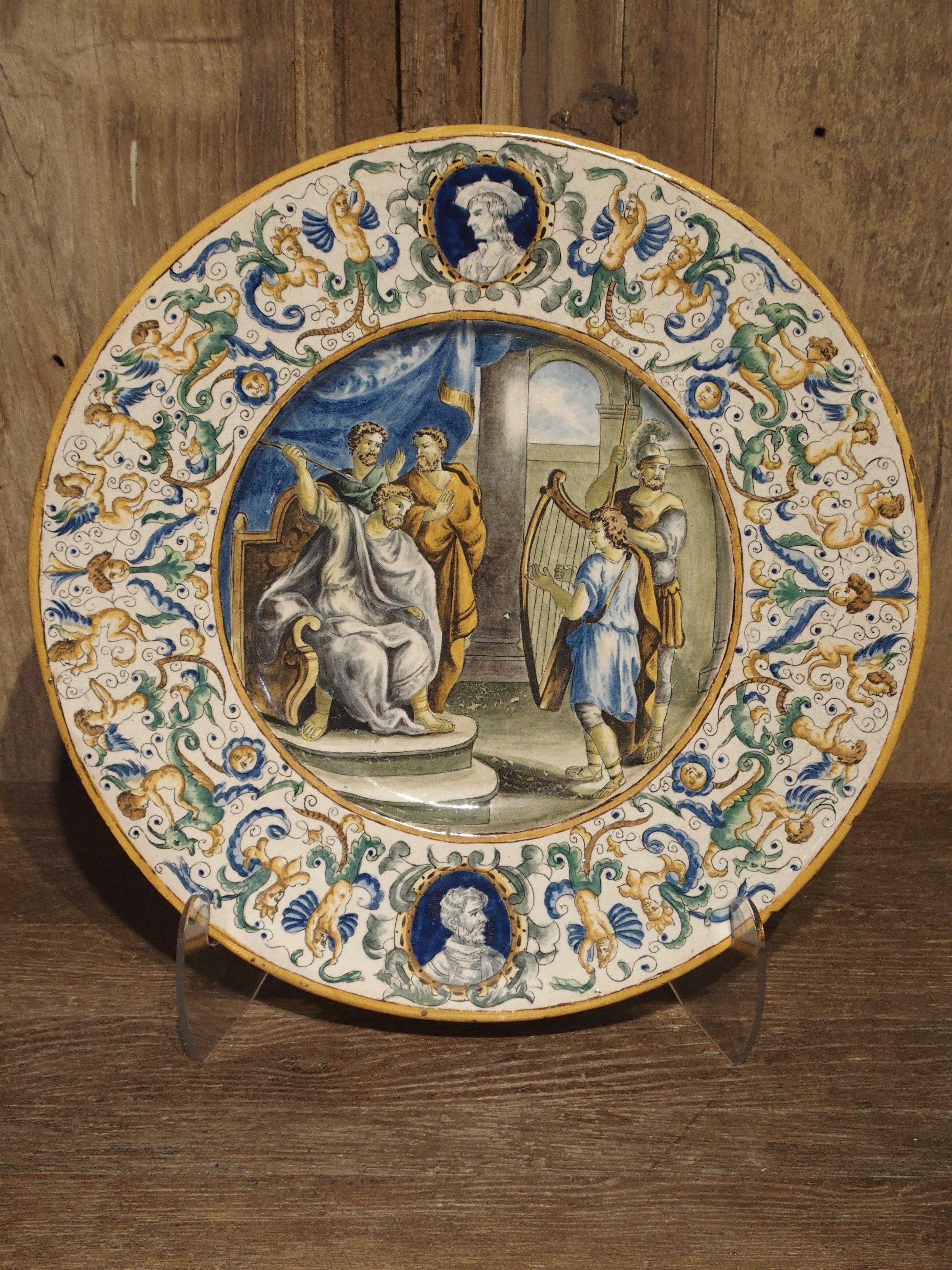 Diese handbemalte Majolika-Platte aus Italien hat zwei Bereiche mit Ornamenten. Der äußere Kreis zeigt die Büsten von zwei verschiedenen Männern, eine oben und eine unten auf der Platte. Diese sind umgeben von einer Fülle von Halbfiguren, Gesichtern