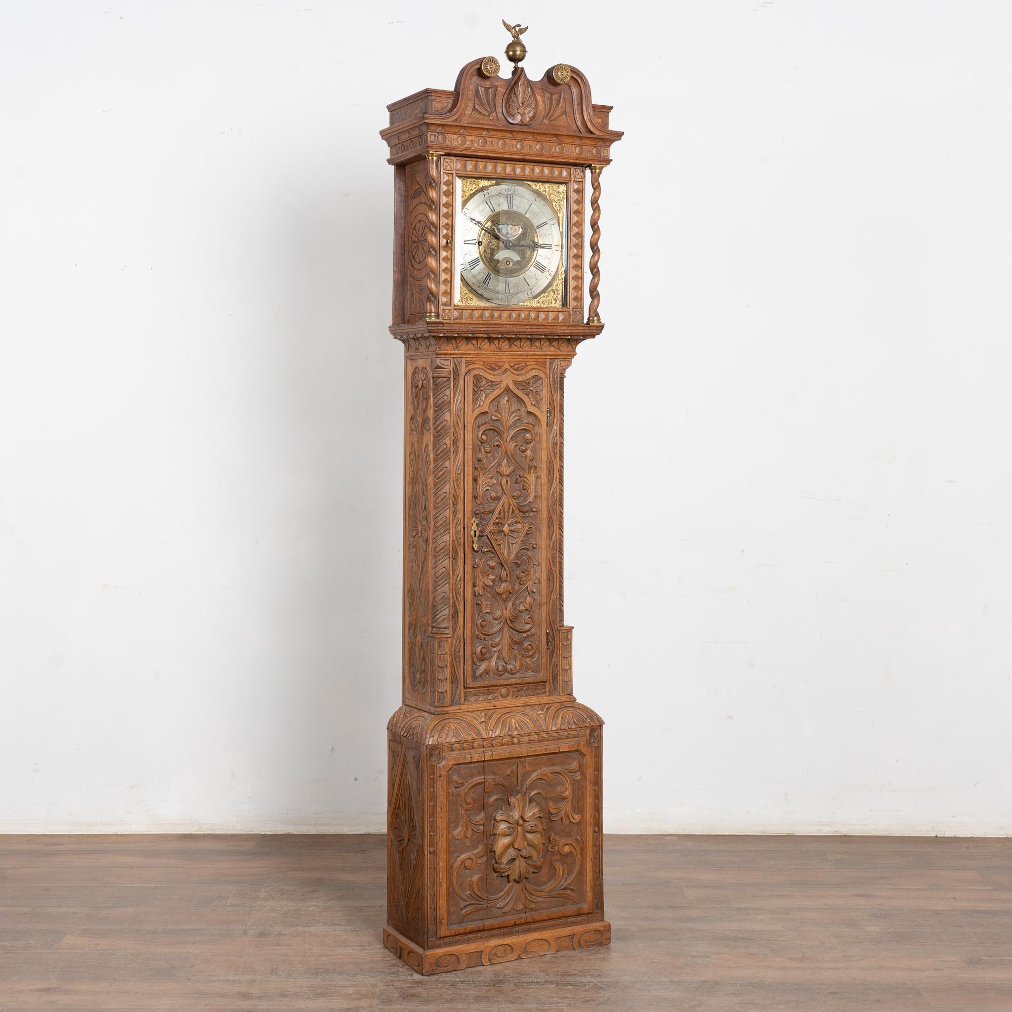 Early 19th-century English oak long-case clock with heavily carved embellishments and rich detail. Turned columns with brass caps frame the beveled glass door and elaborate clock face featuring brass filigree corner accents. An eagle finial crowns