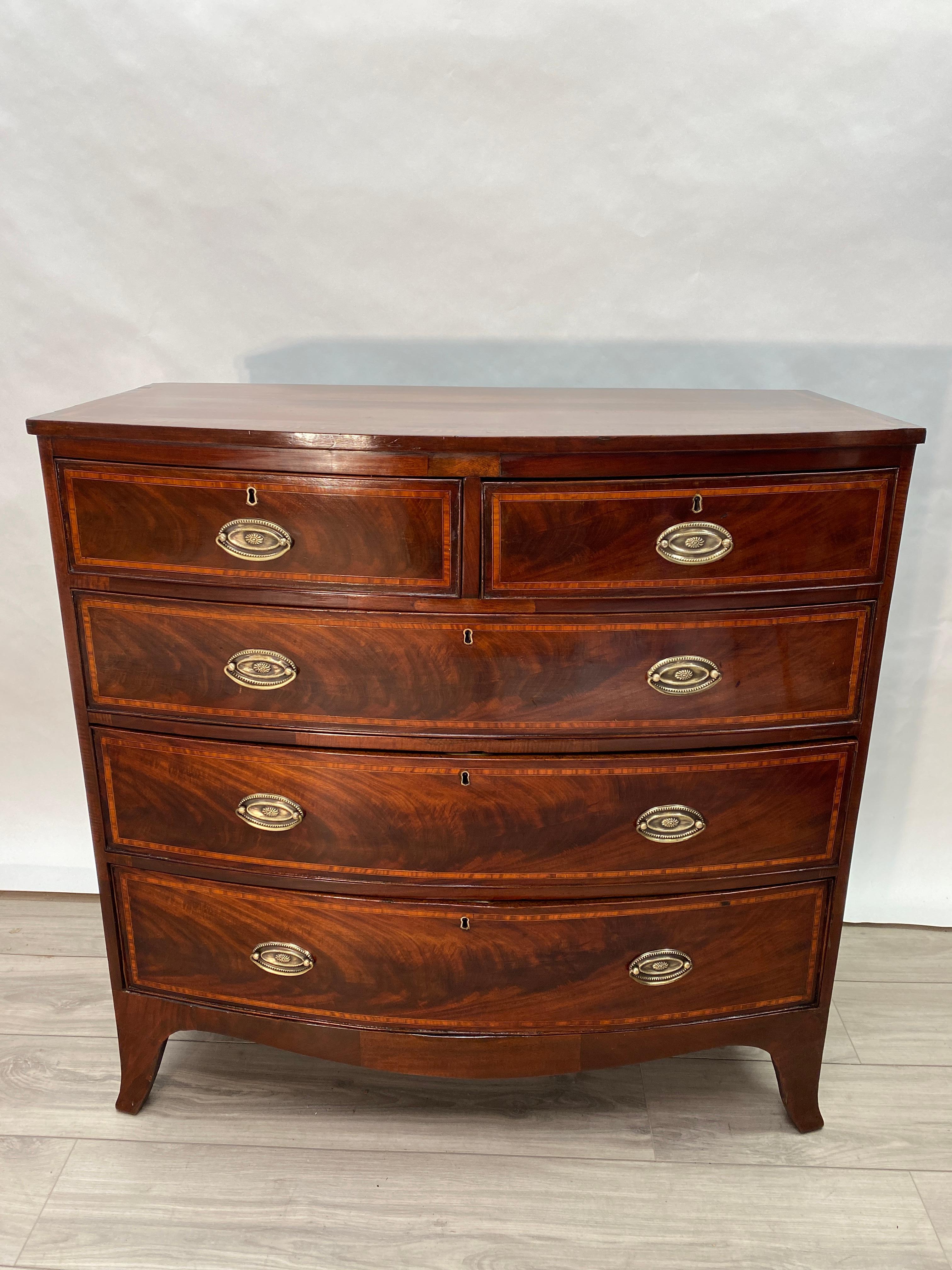 Handsome American made 19th century English Hepplewhite Bow Front chest of drawers. Two over three drawers with bail style brass pulls. Banded veneer encompassing the top. The drawer fronts have been inlayed with flamed mahogany and string banding.
