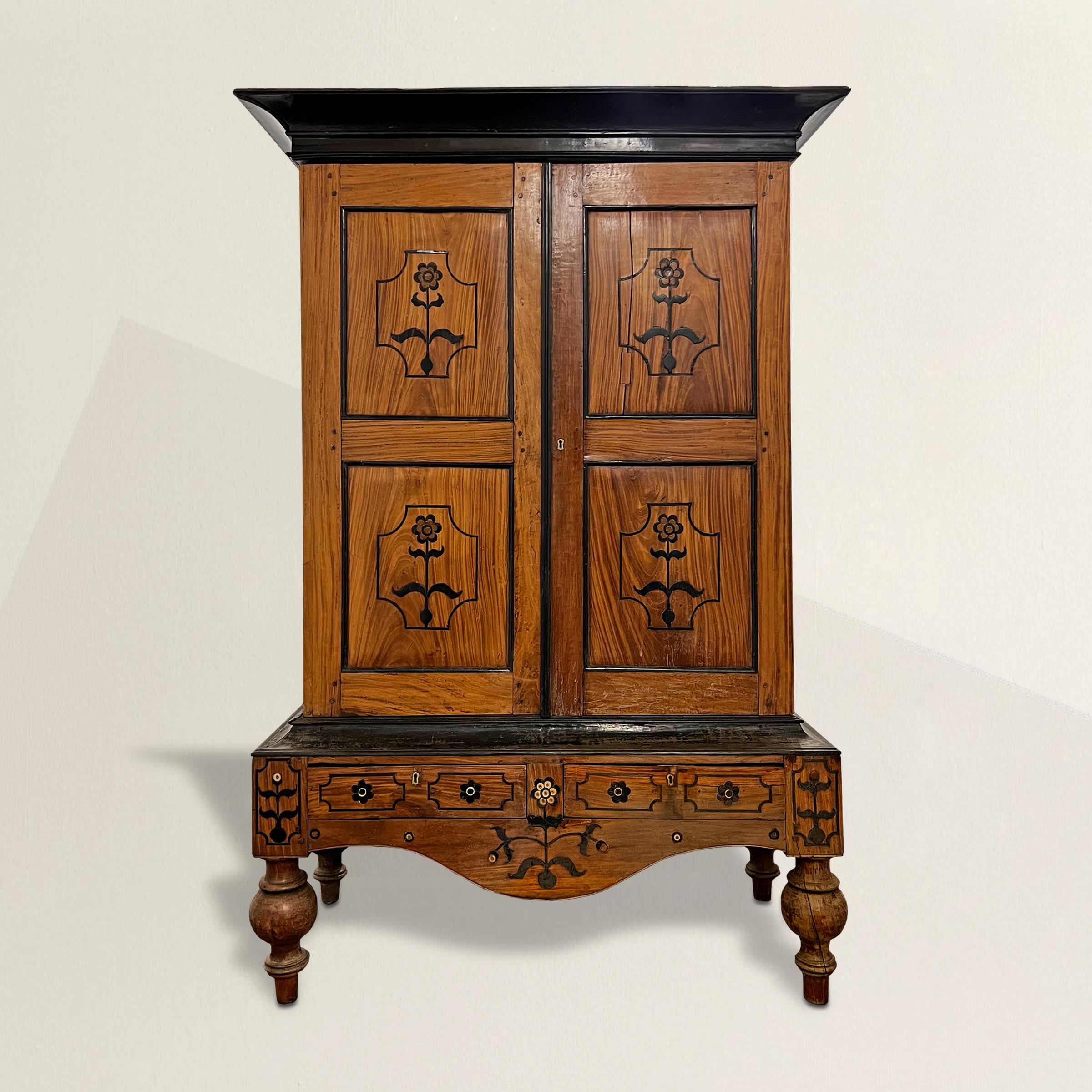 A jaw-dropping and confident 19th century Indian teakwood cabinet with a bold ebonized crown, two doors, each with marquetry inlaid ebonized flowers, a wide apron with two drawers and more inlaid flowers and supported by the most wonderful turned