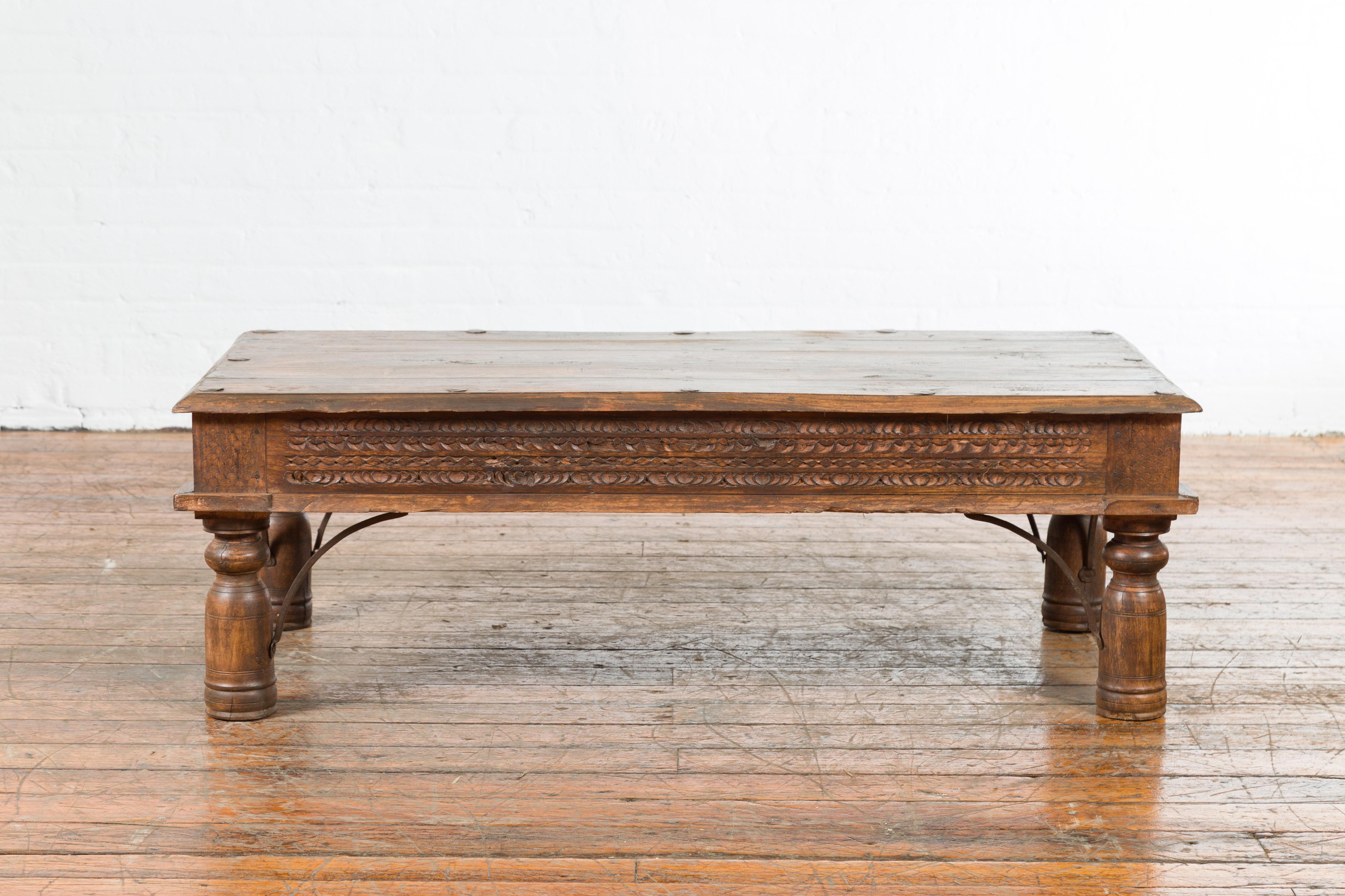 An Indian wooden coffee table from the 19th century, with carved apron, iron accents and baluster legs. Created in India during the 19th century, this coffee table features a rectangular planked top with iron nailheads, sitting above an elegant