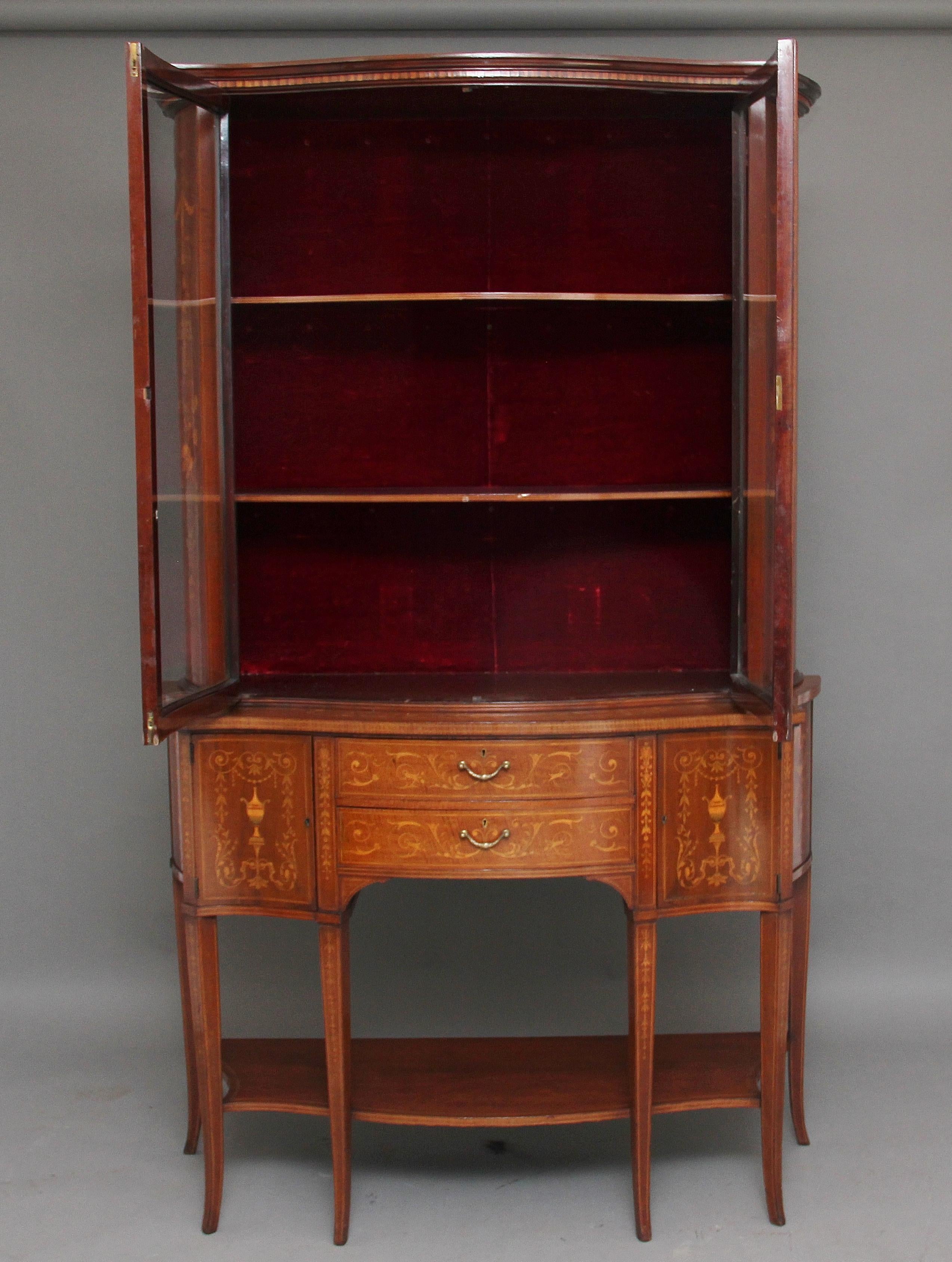 A large 19th century mahogany Sheraton revival inlaid display cabinet of serpentine form, the shaped cornice above two serpentine shaped glazed doors opening to reveal two fixed shelves inside, the sides of the top cabinet profusely decorated with