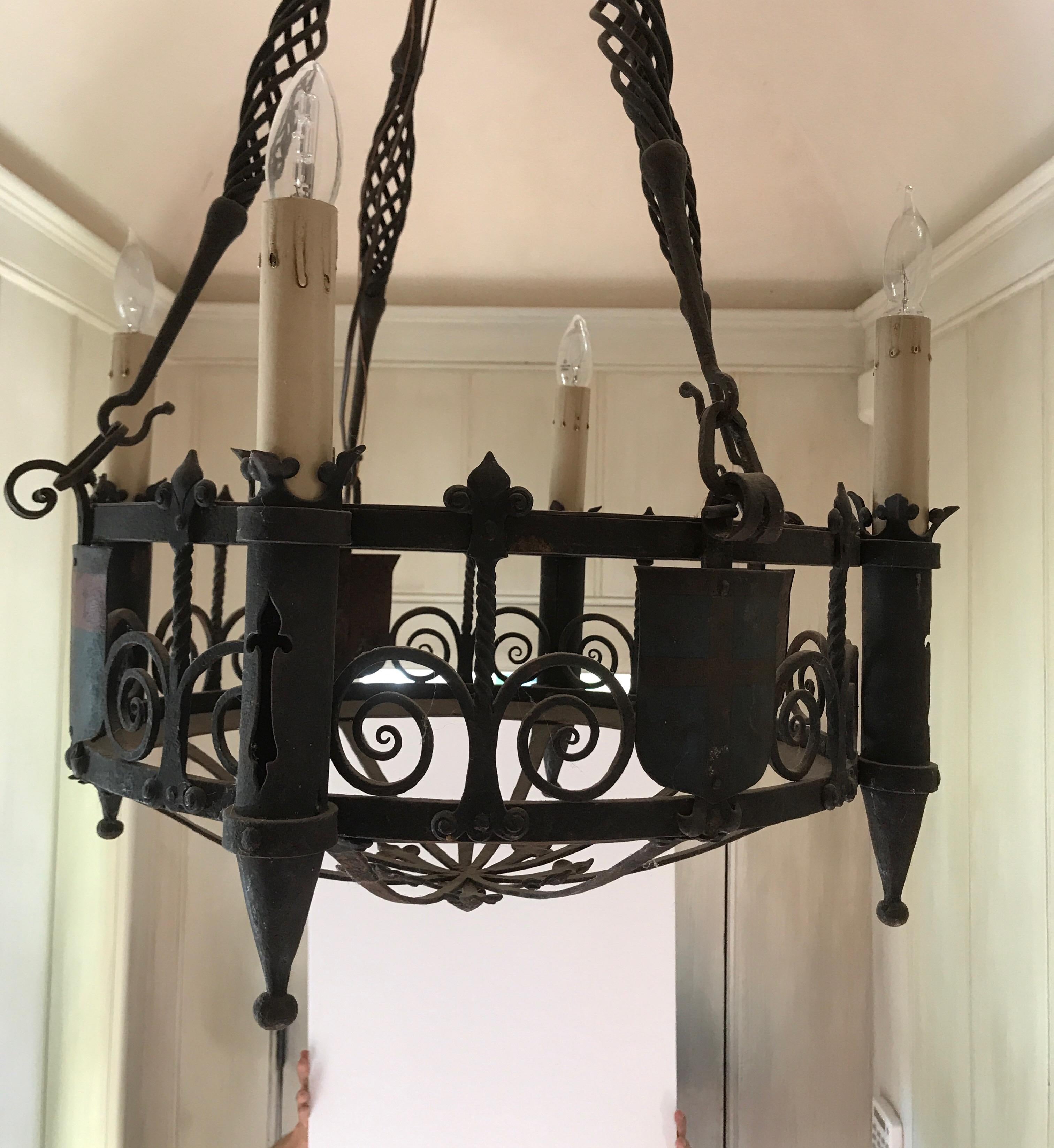 This is a statement iron chandelier with five-lights that are in column form with cut-outs on the candleholders. It is made of beautiful iron work in round iron circles with scroll work in between. Between the candle holders are crest shields