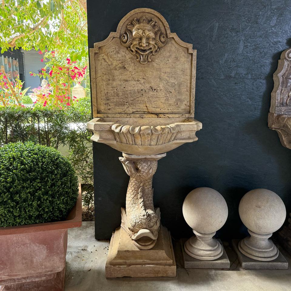 An antique Breccia marble wall fountain. The scalloped water basin is supported by a dolphin sitting on a square base. The backsplash has a very detailed carved mask as the spout. Minor fading, scratches due to age. Wear consistent with age and use.