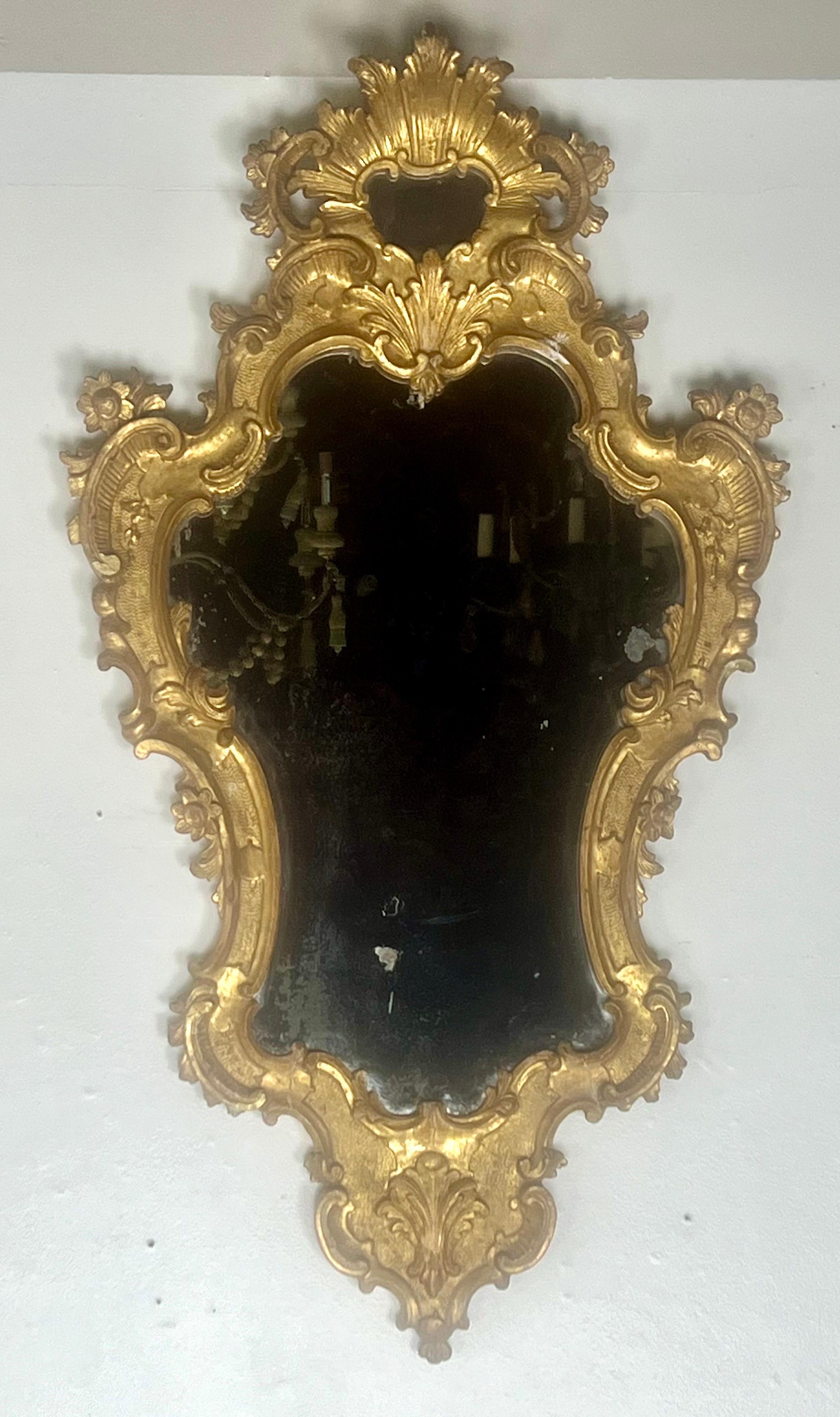 Pair of early 19th-century Rococo style gilt wood mirrors with elaborate frames. The frames have a rich, golden color with intricate details, featuring scrolls, acanthus leaves flowers and shells.  The actual mirror has a distressed finish and has