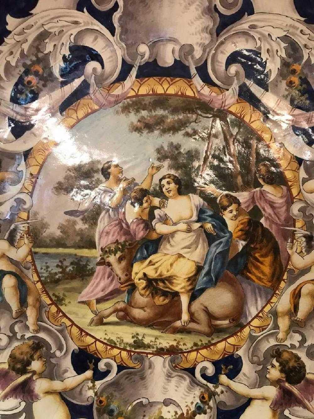 Crafted of earthenware pottery having a hand painted center landscape scene with guardians and bovine. The border depicts winged putti, floral garlands, stylized shells, and leaf scrolls. Colorfully decorated in tin glaze. Excellent condition. 25