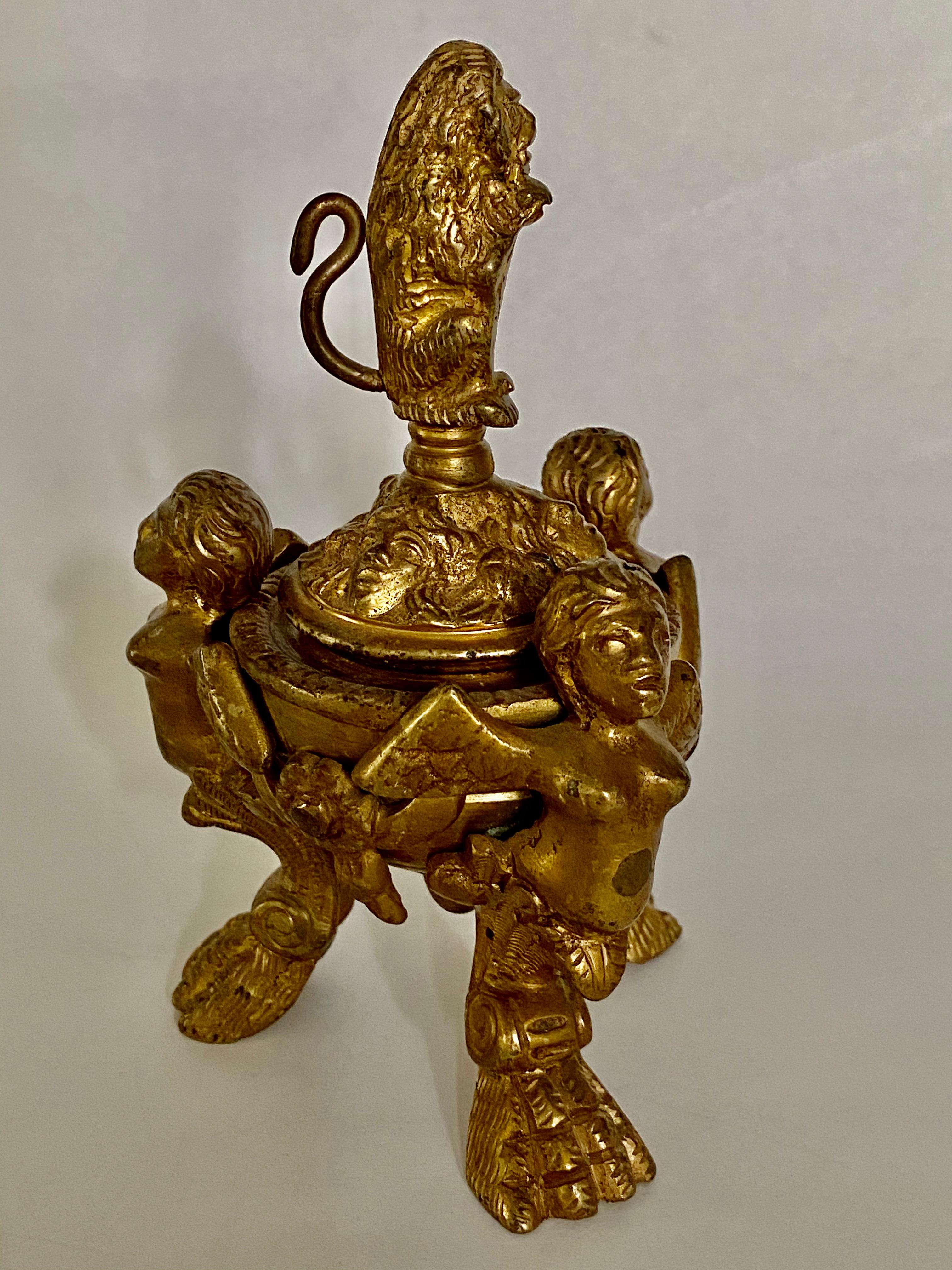 After a 16th century Venetian design, winged putti with taloned legs support a turned bronze inkwell covered with a lid decorated with masks and garlands, surmounted by a lion holding a shield.