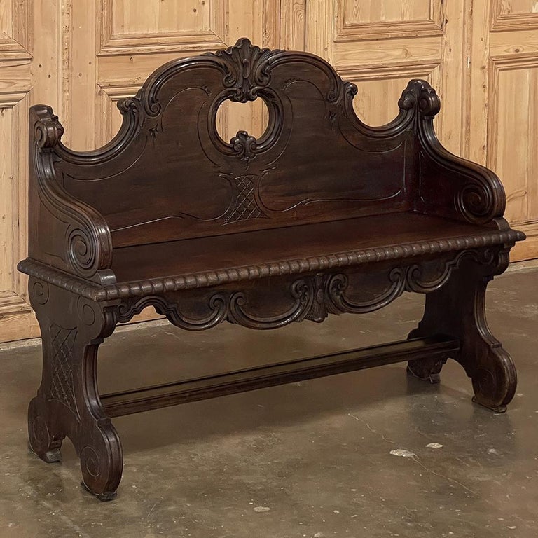 19th Century Italian hall bench was sculpted from fine walnut, and features an elaborately scrolled design including a boldly arched seatback embellished with foliate scrollwork and centered with a stylized shell shield motif appearing over an