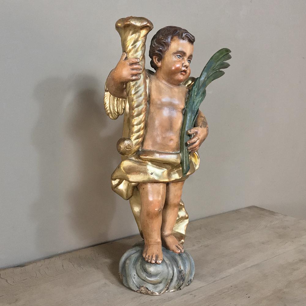 19th century Italian hand-carved and painted cherub presents a charming, colorful and adorable rendering of a young winged cherub, holding a spiral torchere in one hand that can be used for a candle, and a palm frond in the other. Vivid coloration