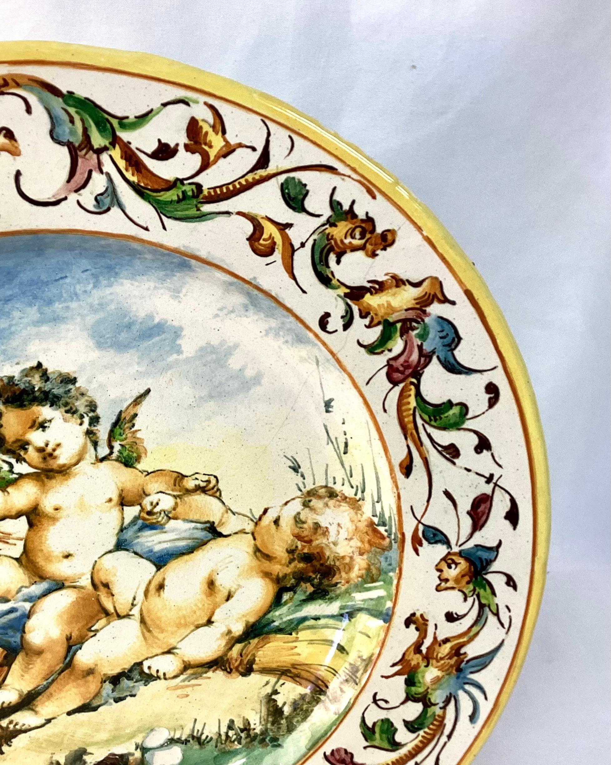 19th century hand painted Italian charger featuring three cherubs interacting with another lying on a bed of wheat stalks under a scenic skyscape. Beautiful yellow, blue and green color palette.
