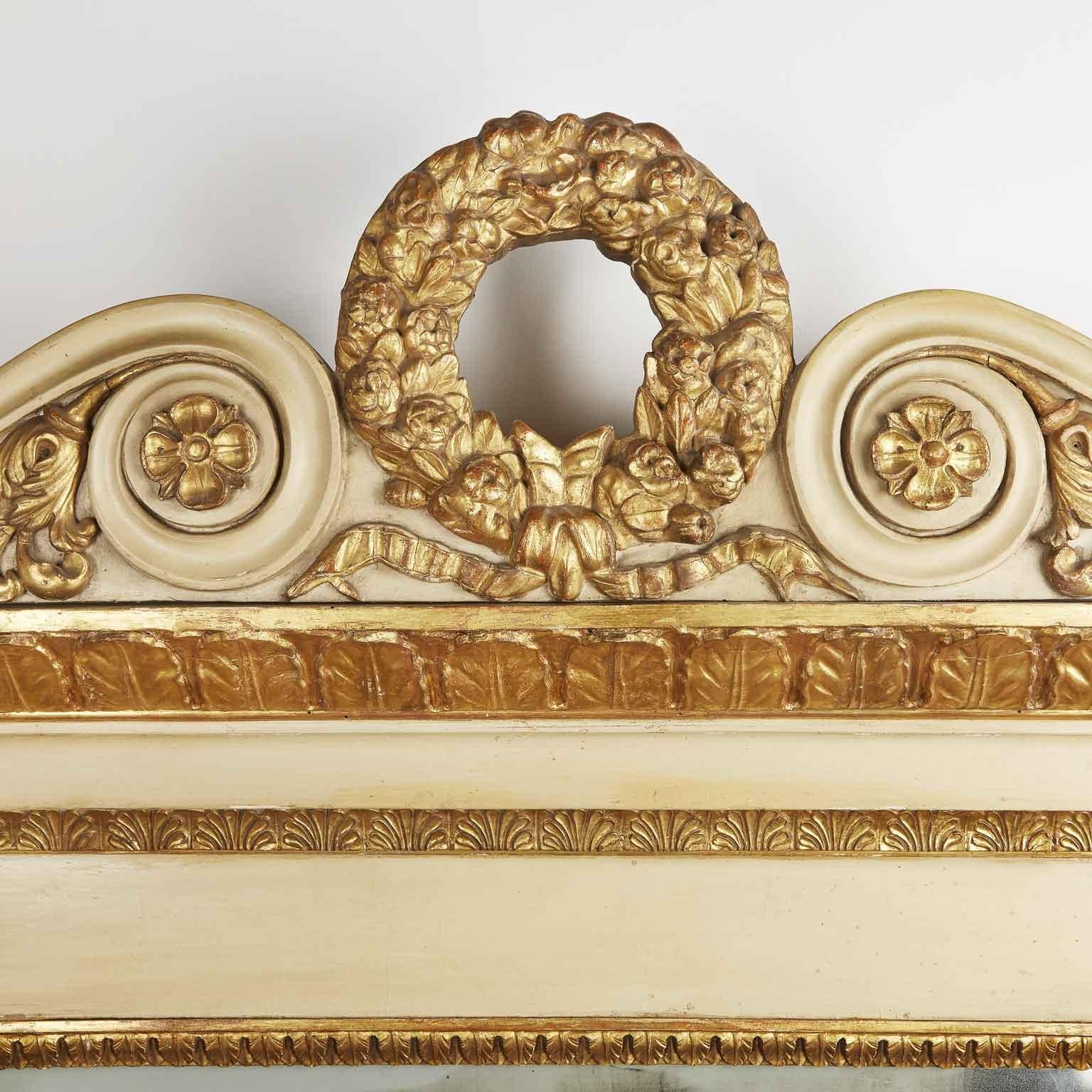 Italian antique ivory color painted and giltwood fireplace mirror, dating back to the mid-19th century. This elegant Neoclassical overmantel mirror is decorated with carved giltwood floral and scrolling ornament, centered by a circular garland. The