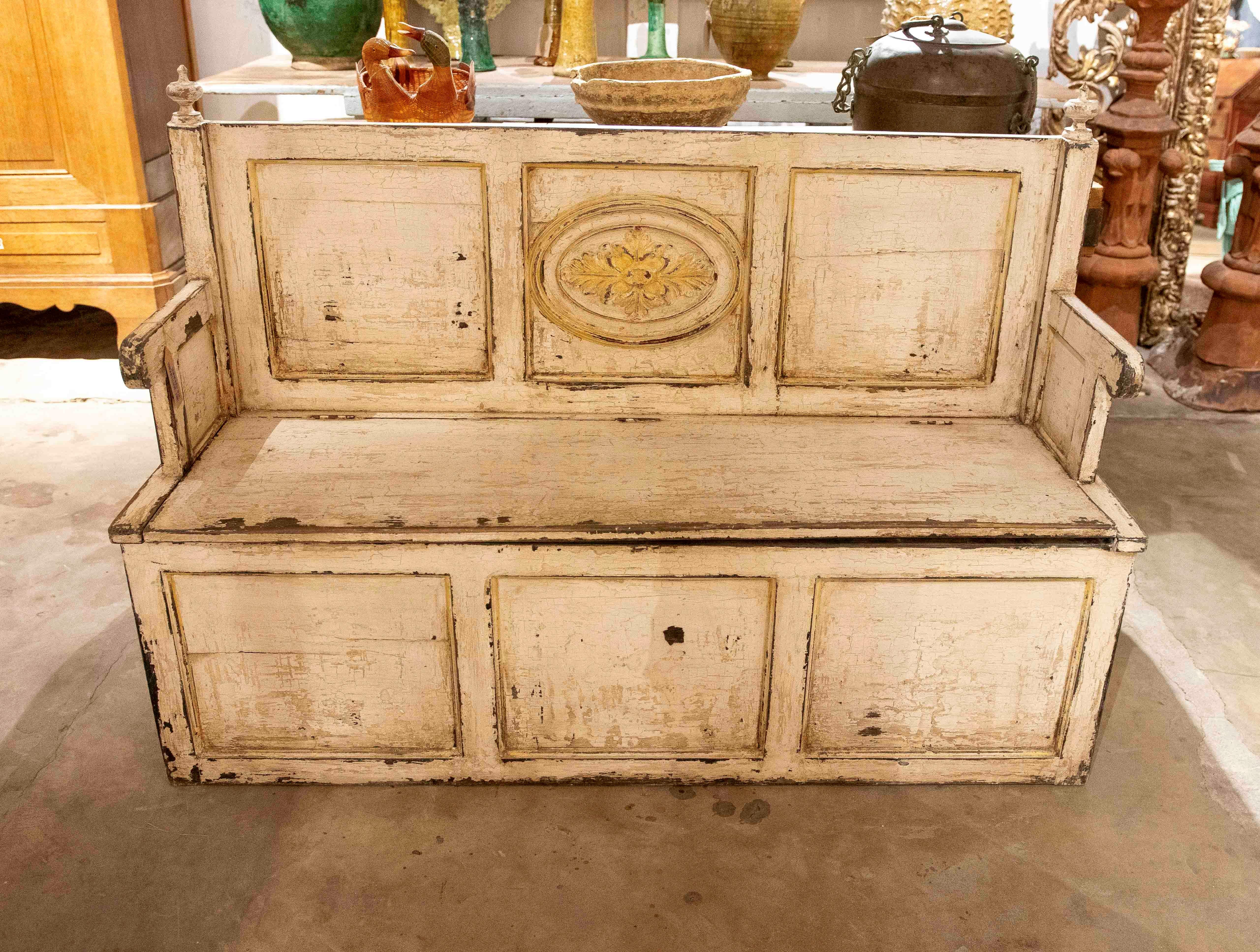 Charming vintage storage bench, made of solid wood and painted in a creamy tone with an original patina and wear that reveals its age. The backrest is decorated with molded panels and a carved floral medallion in the center, adding elegance and