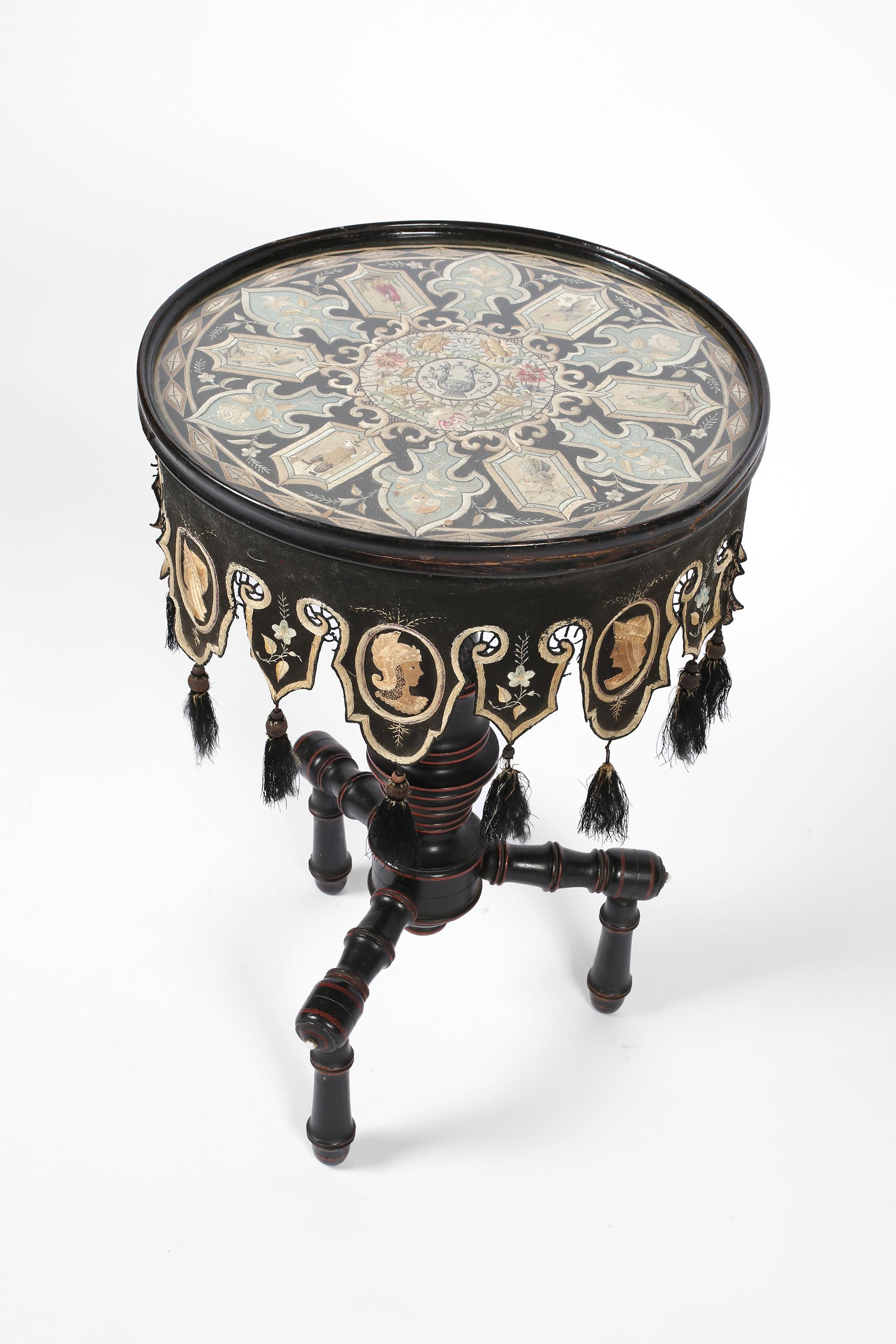 Un insolito tavolo occasionale del XIX secolo in legno tornito, ebanizzato e con sfumature cremisi. Protetto da un vetro, l'intricato top ricamato in seta e le frange con nappe, raffigurano figure che suonano strumenti, uccelli tropicali e busti in