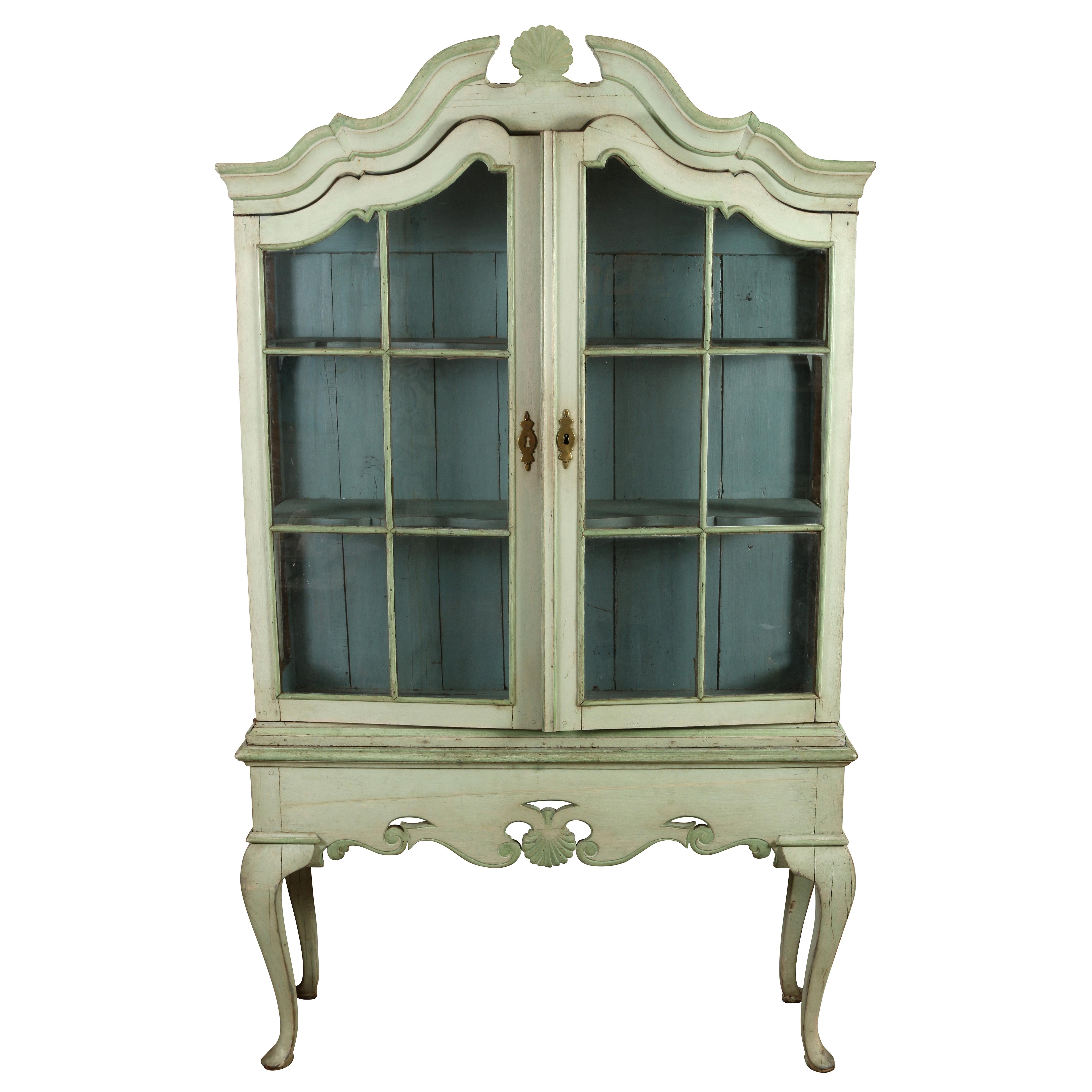 We love the charm and whimsy of this special vitrine cabinet.  Painted in a light green with a deep blue interior, the top  portion has glazed doors with the original glass,  The broken pediment and the apron on lower section are decorated with a