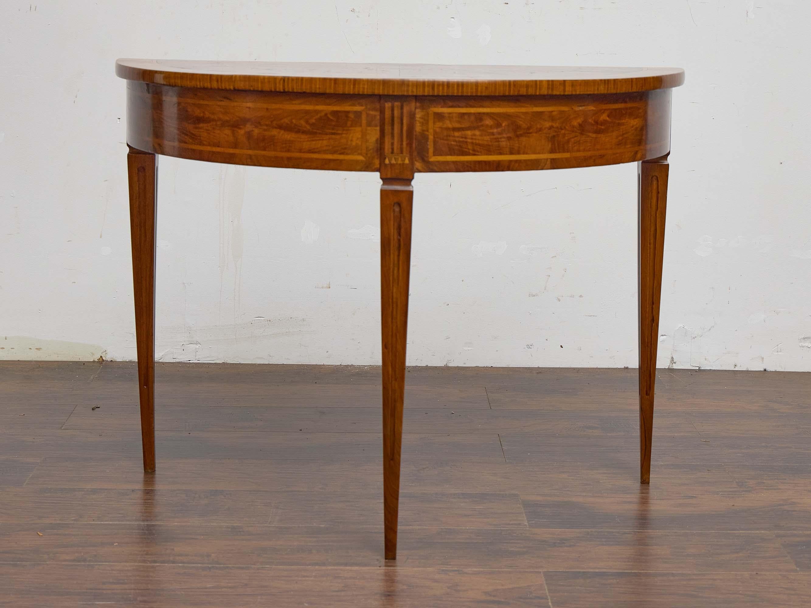 An elegant Italian walnut demi-lune table from the 19th century, supported on three tapered and fluted legs that emphasize its neoclassical character. The half-moon top is veneered in walnut with a radiating design, framed by a satinwood banded
