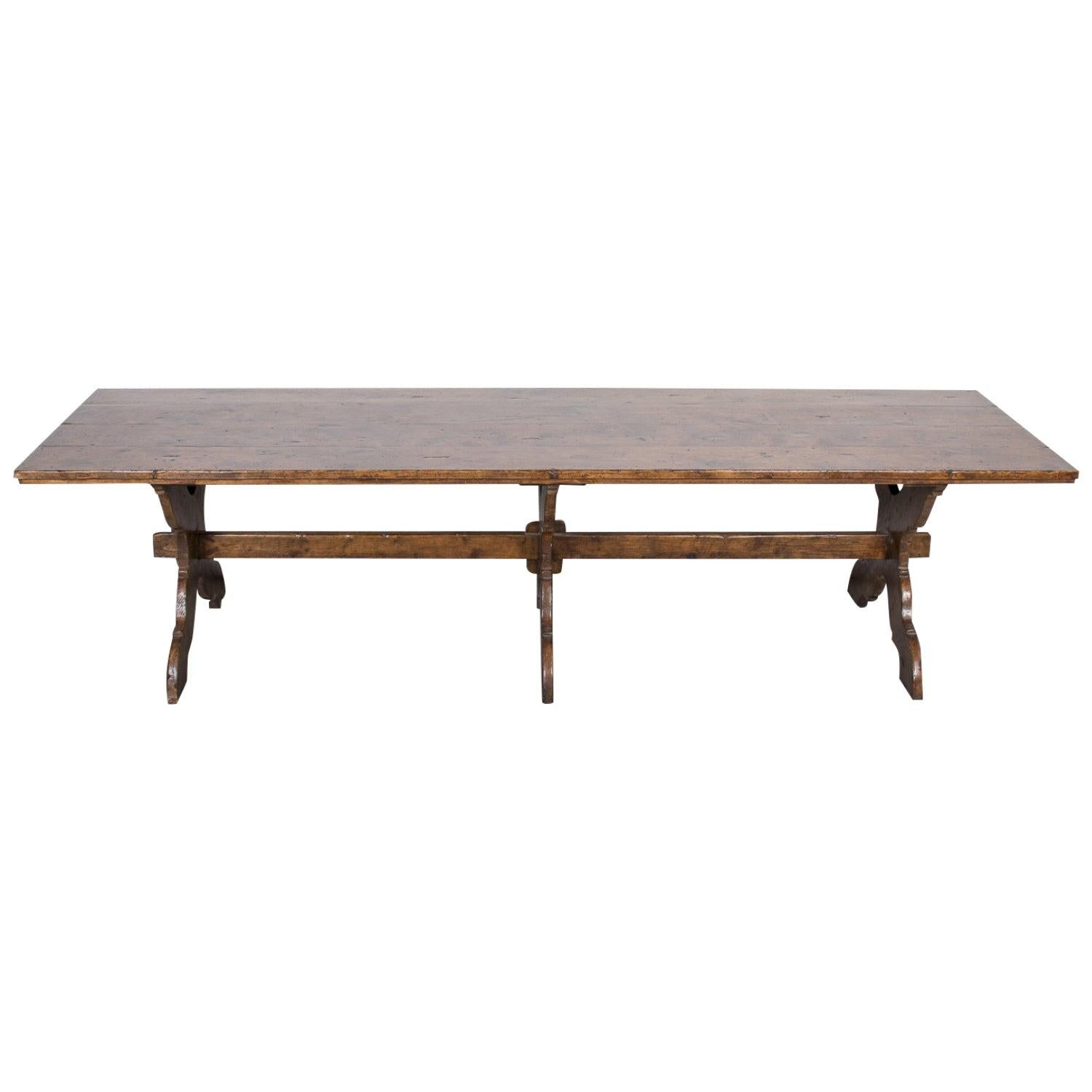 Fabriquée à la main à la fin du XIXe siècle par des artisans talentueux dans les collines ensoleillées de la Toscane, cette monumentale table à tréteaux ou table de récolte en noyer italien sculpté présente un épais plateau rectangulaire en noyer