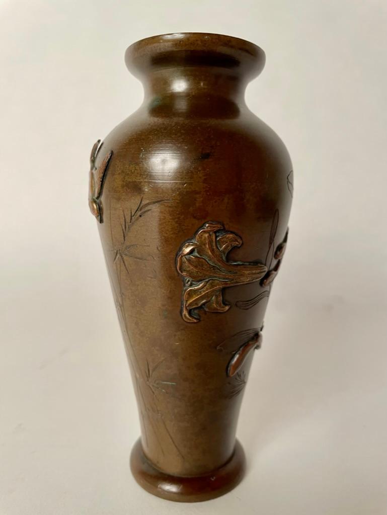 petit vase en bronze de la période Meiji, datant du XIXe siècle, avec une belle décoration gravée et appliquée. Un oiseau doré en relief vole au-dessus de bambous gravés d'un côté, et des lys appliqués dorés avec des feuilles patinées de l'autre. Un