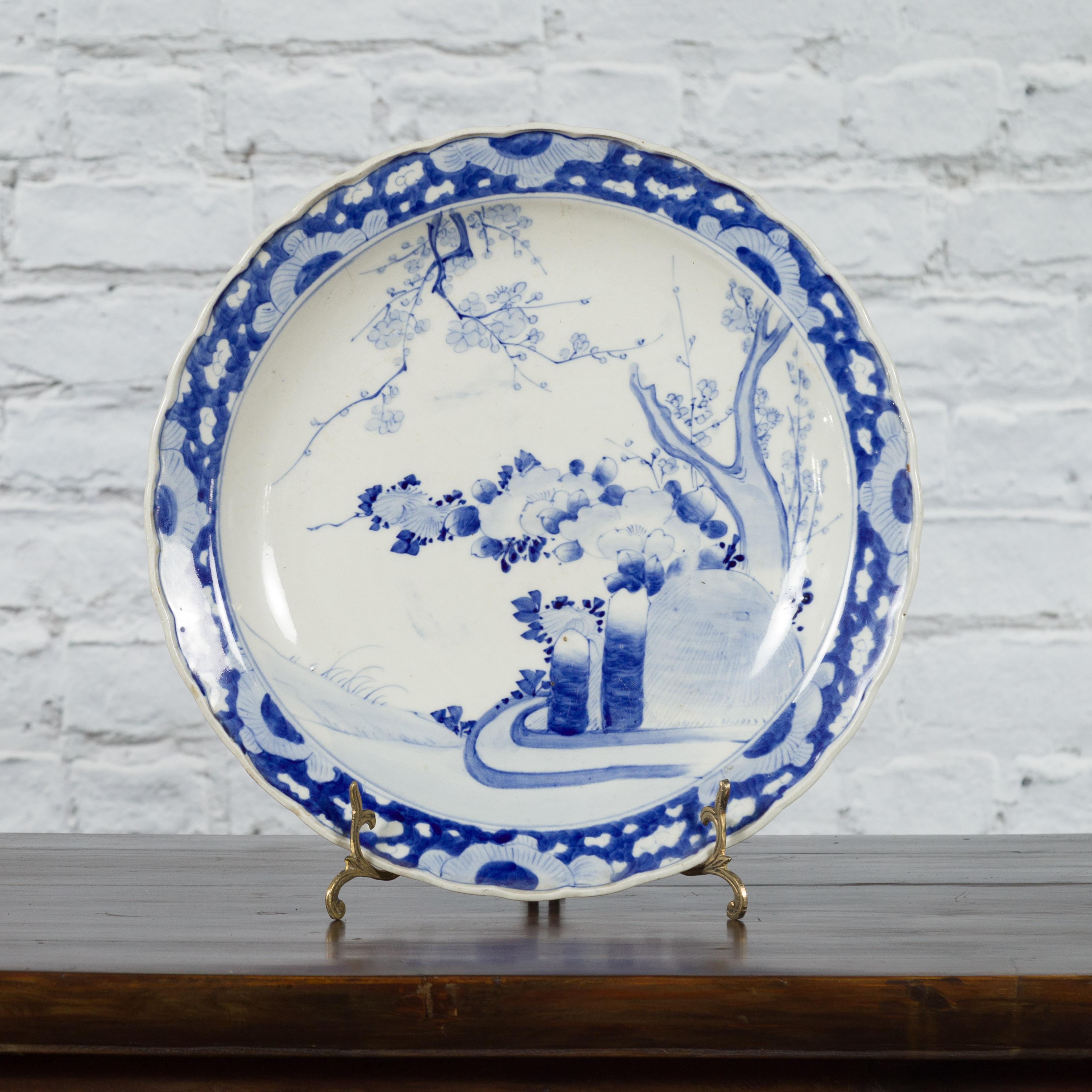 A Japanese hand-painted porcelain plate from the 19th century, with blue and white tree and foliage décor and scalloped edge. Created in Japan during the 19th century, this porcelain plate features a delicate blue and white décor depicting a