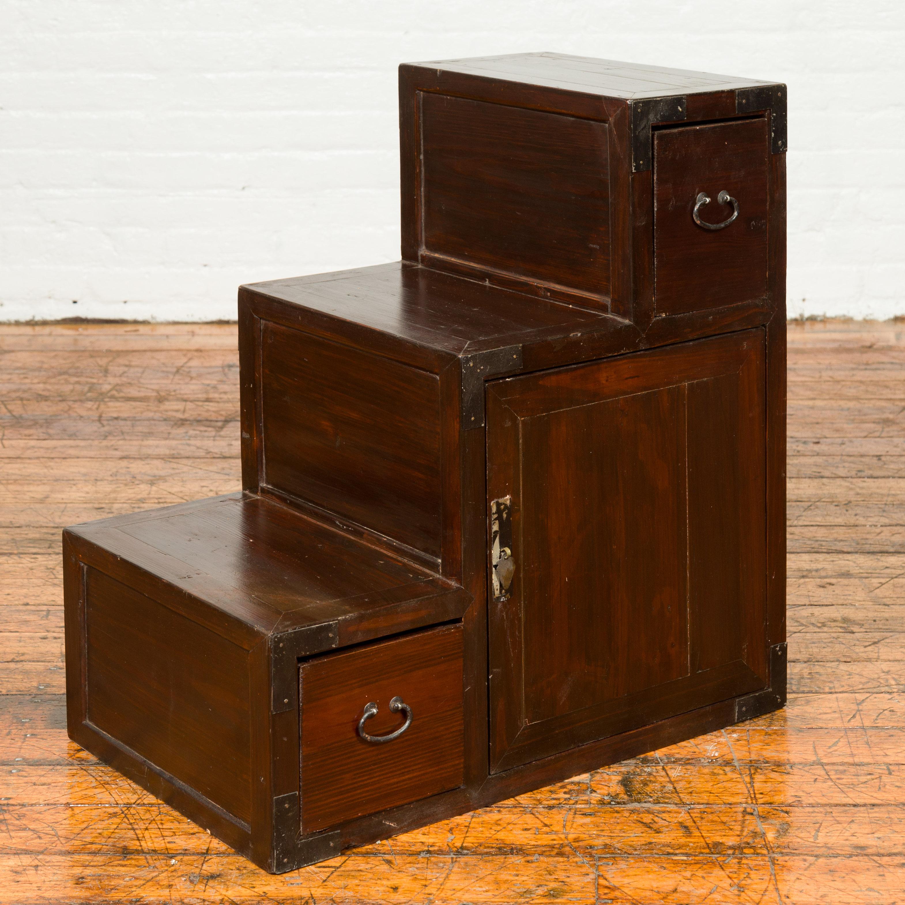 A Japanese staircase tansu cabinet from the 19th century, with two drawers and single door. Born in Japan during the 19th century, this staircase tansu features a convenient stepped storage unit presenting a dark patina. Ideal to display decorative