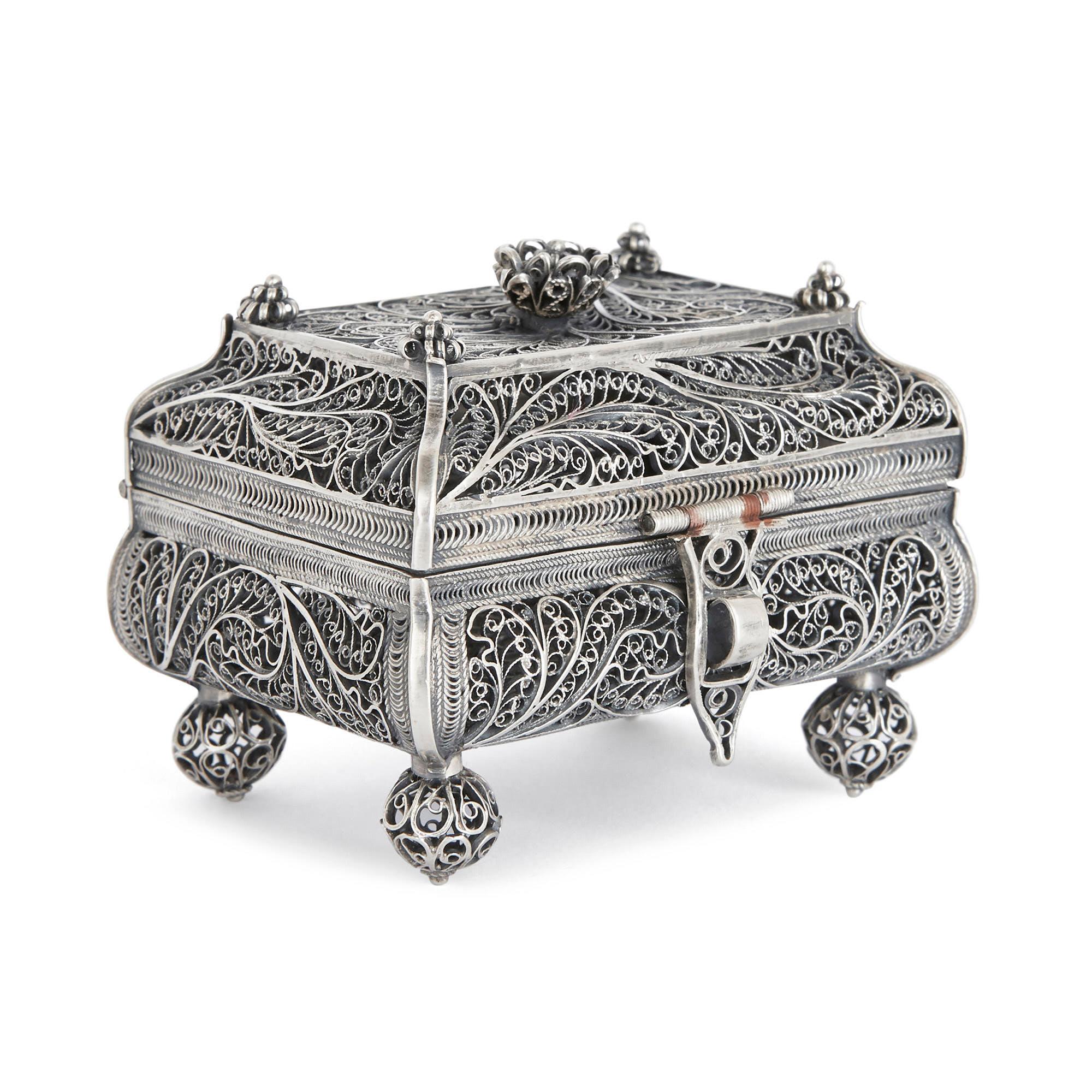 The silver box is set on four ball feet. Its body is of rectangular form, with curved edges, and it is covered by a shaped lid with a flat top. The lid is shut by a decorative clasp. Its top corners are decorated with small flower finials, and its