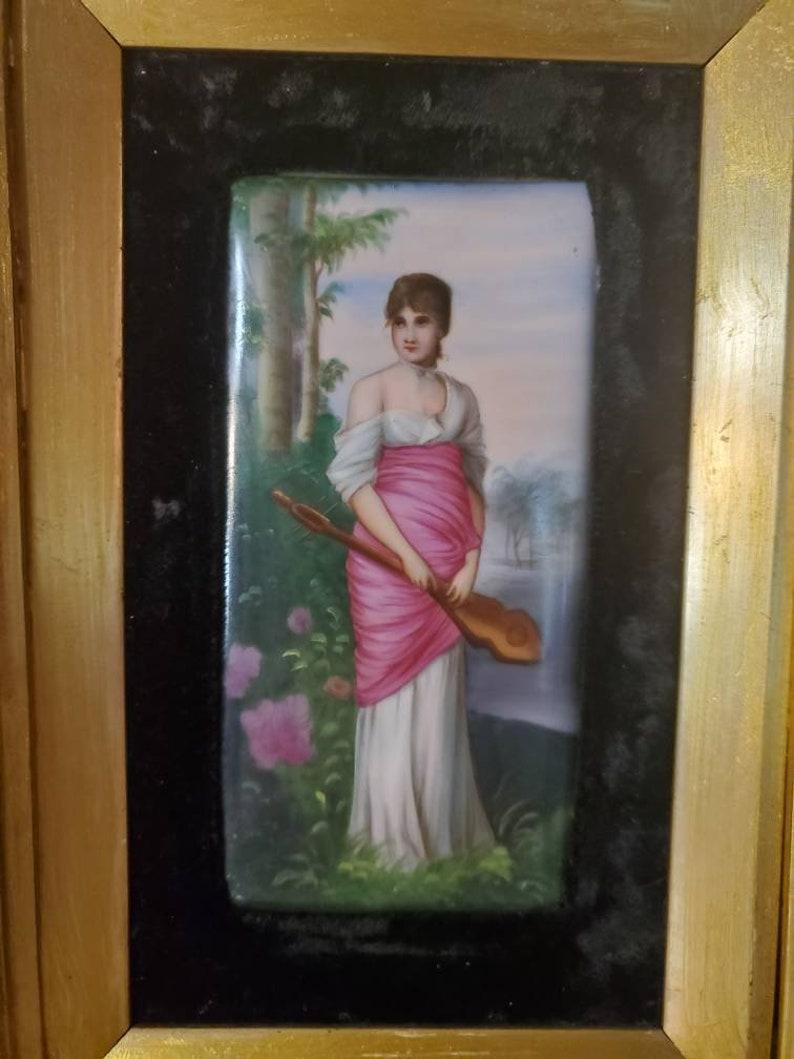 A late 19th century Berlin KPM painted porcelain plaque, Die Lautenspielerin, in pierced foliate giltwood frame, painting after Friedrich August von Kaulbach (German, 1850-1920). Modeled as a standing auburn-haired maiden holding a lute in a wooded