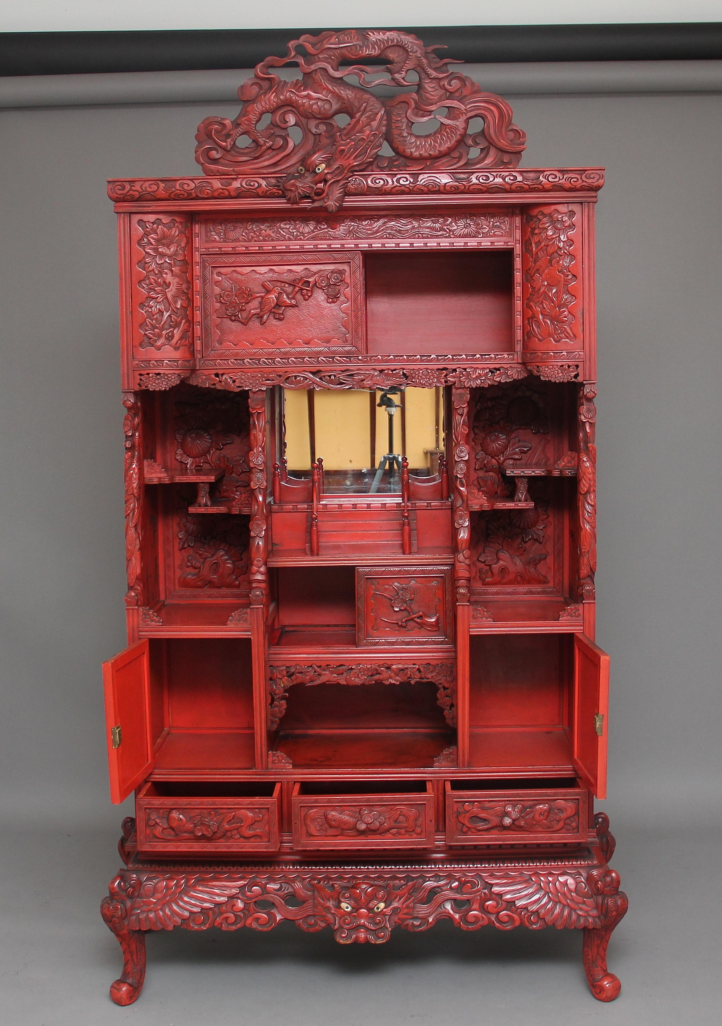 A highly decorative 19th century red lacquered Japanese shodona cabinet, having a large dragon pediment above the cabinet with the head looking over the carved cornice, the cabinet consisting of various shelves, hinged and sliding doors, with three