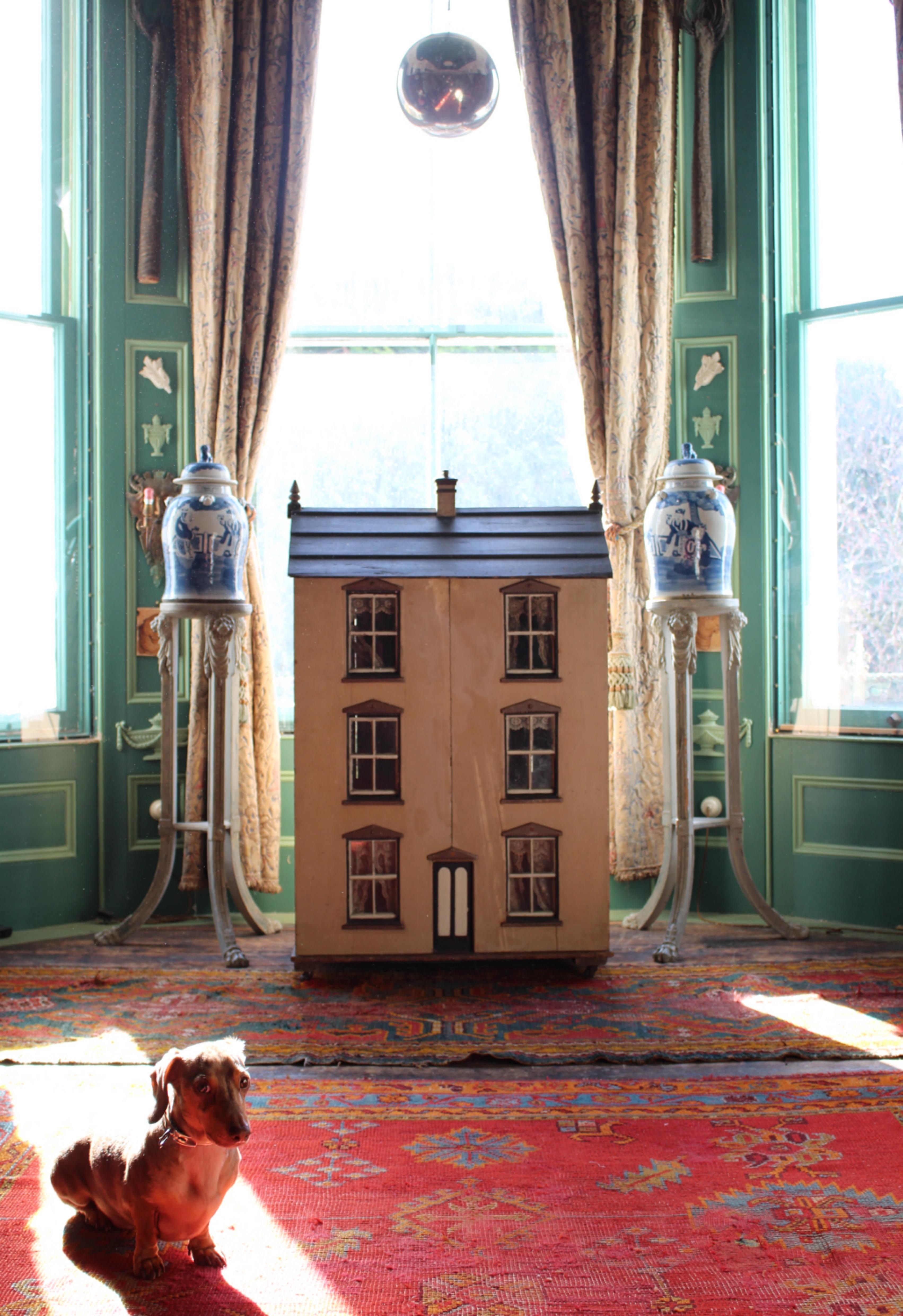A large and well proportioned three storey dolls house with a peak roof and central chimney over a six sash window facade and centre door.

The house has its original painted cream exterior and black clad roof, the back with hinged doors for access