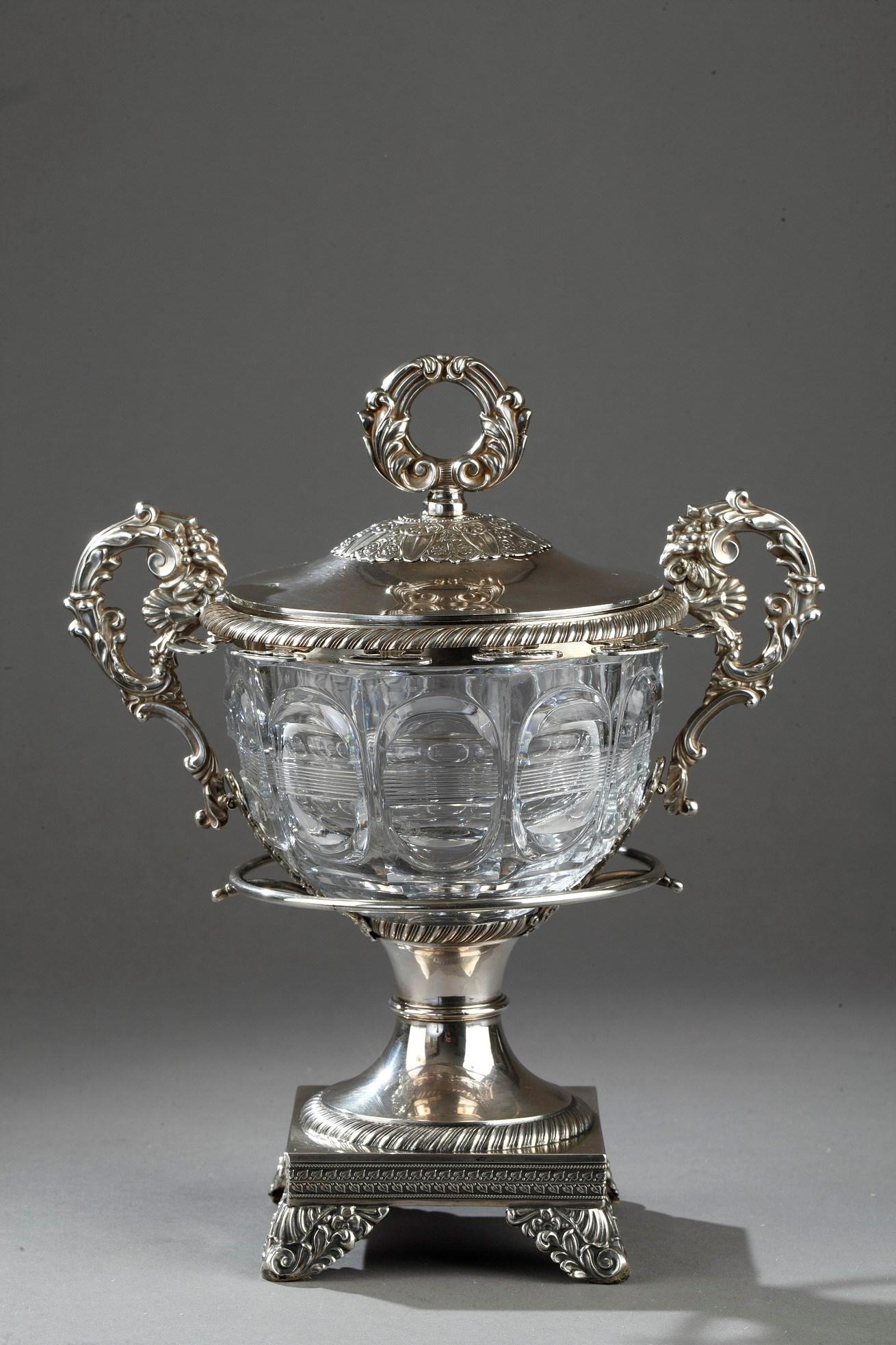 Silver and cut-crystal confiturier decorated with geometrical patterns.The silver frame is adorned with laurel leaves and stylized palmettes. The handles are decorated with acanthus leaves, seeds and shells forming a cornucopia. Twelve vermeil