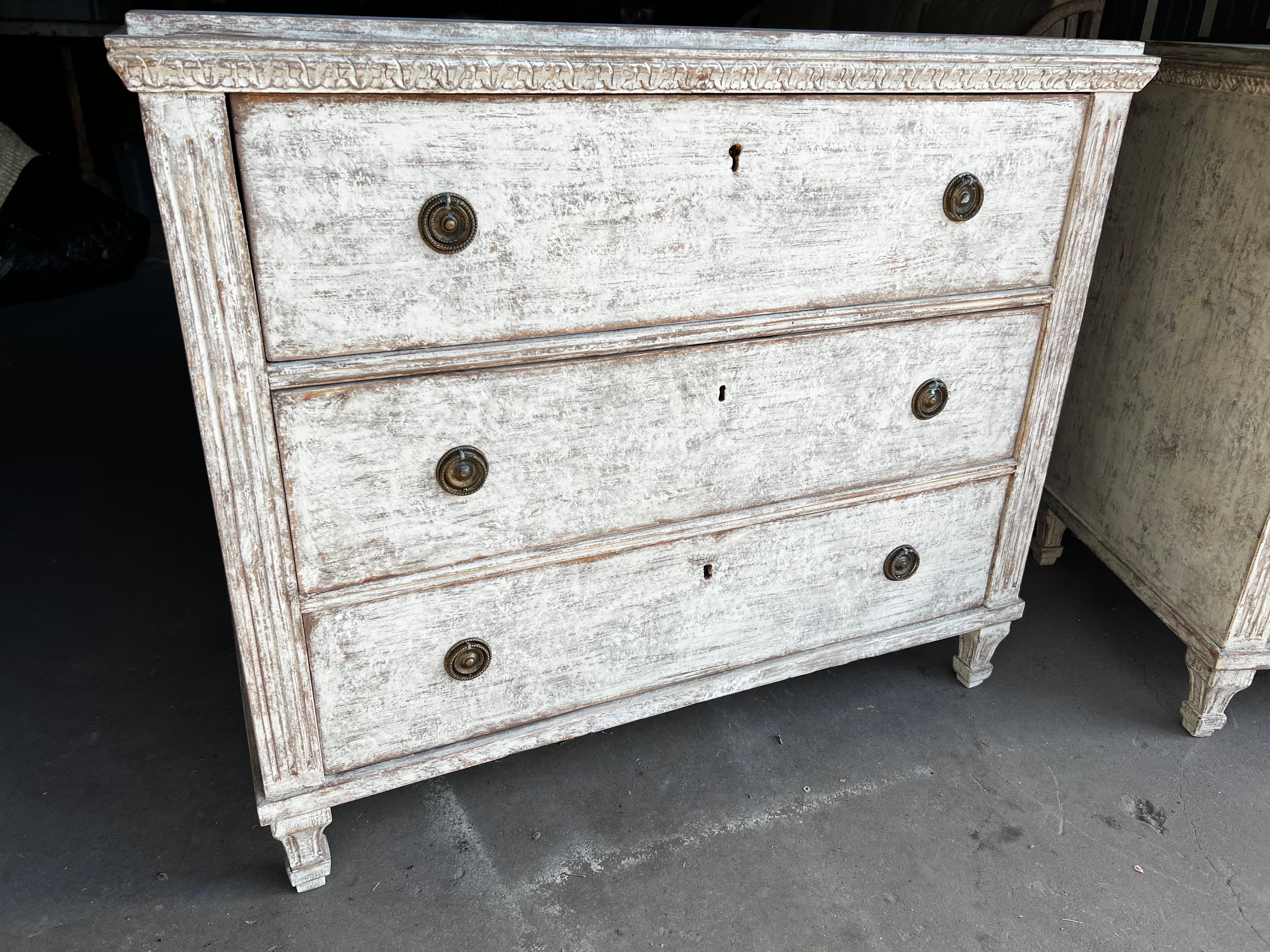 A pair of stunning 19th century Neoclassical Swedish Gustavian style painted wood commodes. This gorgeous pair features three drawers with classical brass hardware with fluted side posts that are resting on short tapered, fluted feet. Painted in a