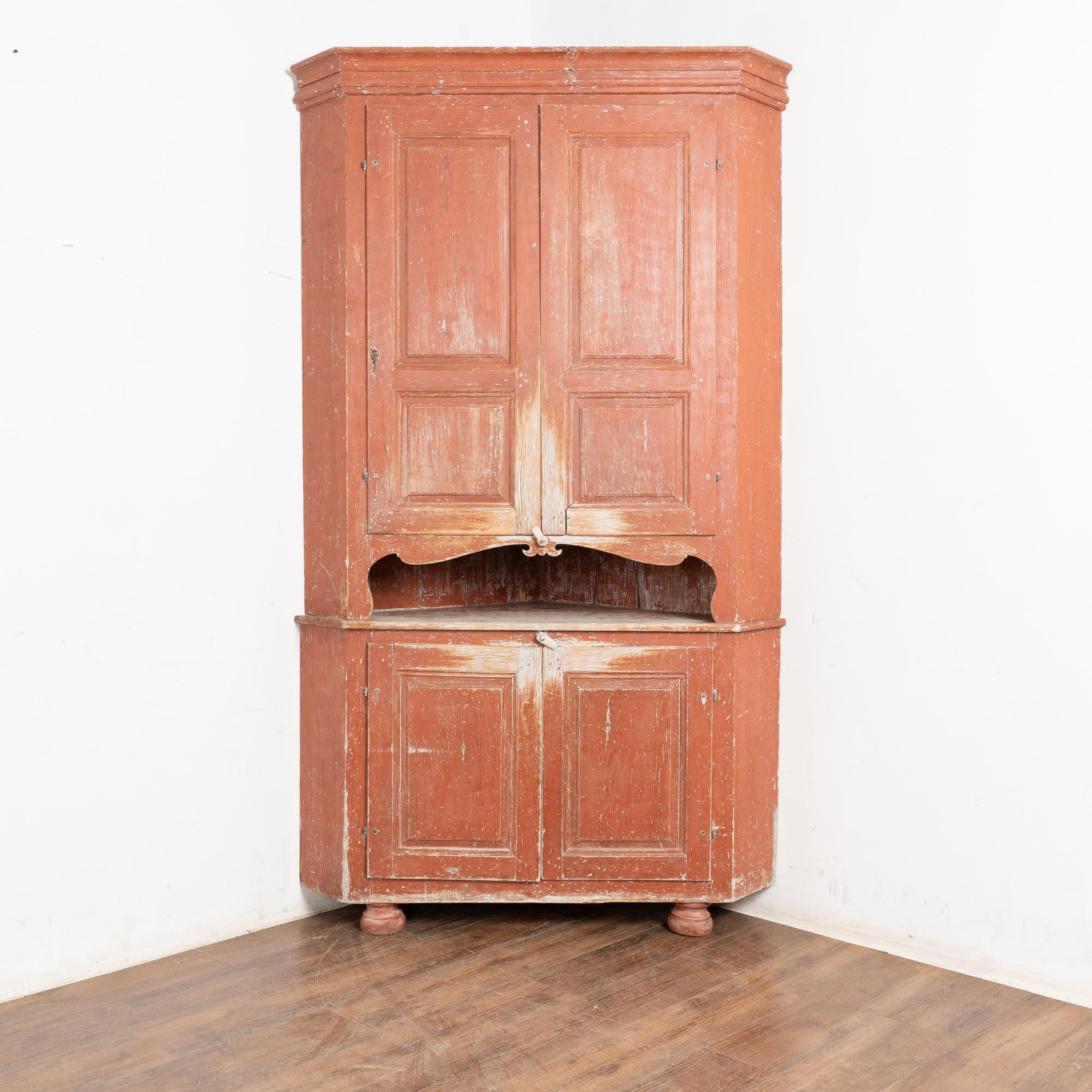 This large pine corner cabinet still maintains the original brick red painted finish, now worn down through generations of use to the natural pine below. Specks of white and white-washed pine also visible. 
It is built in two sections and reaches
