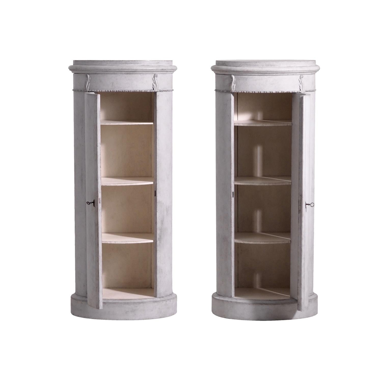A light-grey, antique Swedish Gustavian pair of pedestal cabinets with four shelfs made of hand crafted painted Pinewood, standing on a round base, enhanced by detailed wood carvings, in good condition. The Scandinavian pair are particularized in