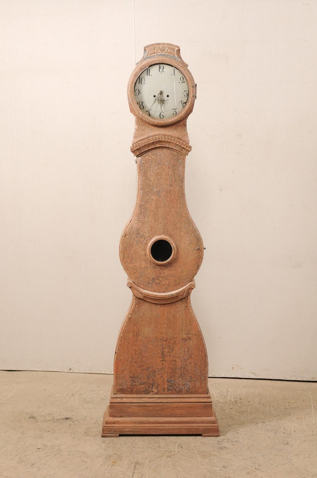 A 19th century Swedish long-case clock with dry-scraped finish. This antique clock from Sweden, circa 1820s, is topped with a subtly raised and carved crest, atop a round-shaped head. A nicely trimmed molding accents the neck, just above the