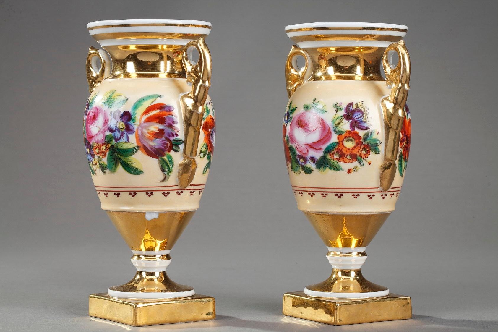 Pair of small gilded and white porcelain vases with an Etruscan shape. The body of the vases is in light ochre, and decorated with a highly detailed frieze of bright, polychromatic flowers. These centerpiece vases feature short handles accented with
