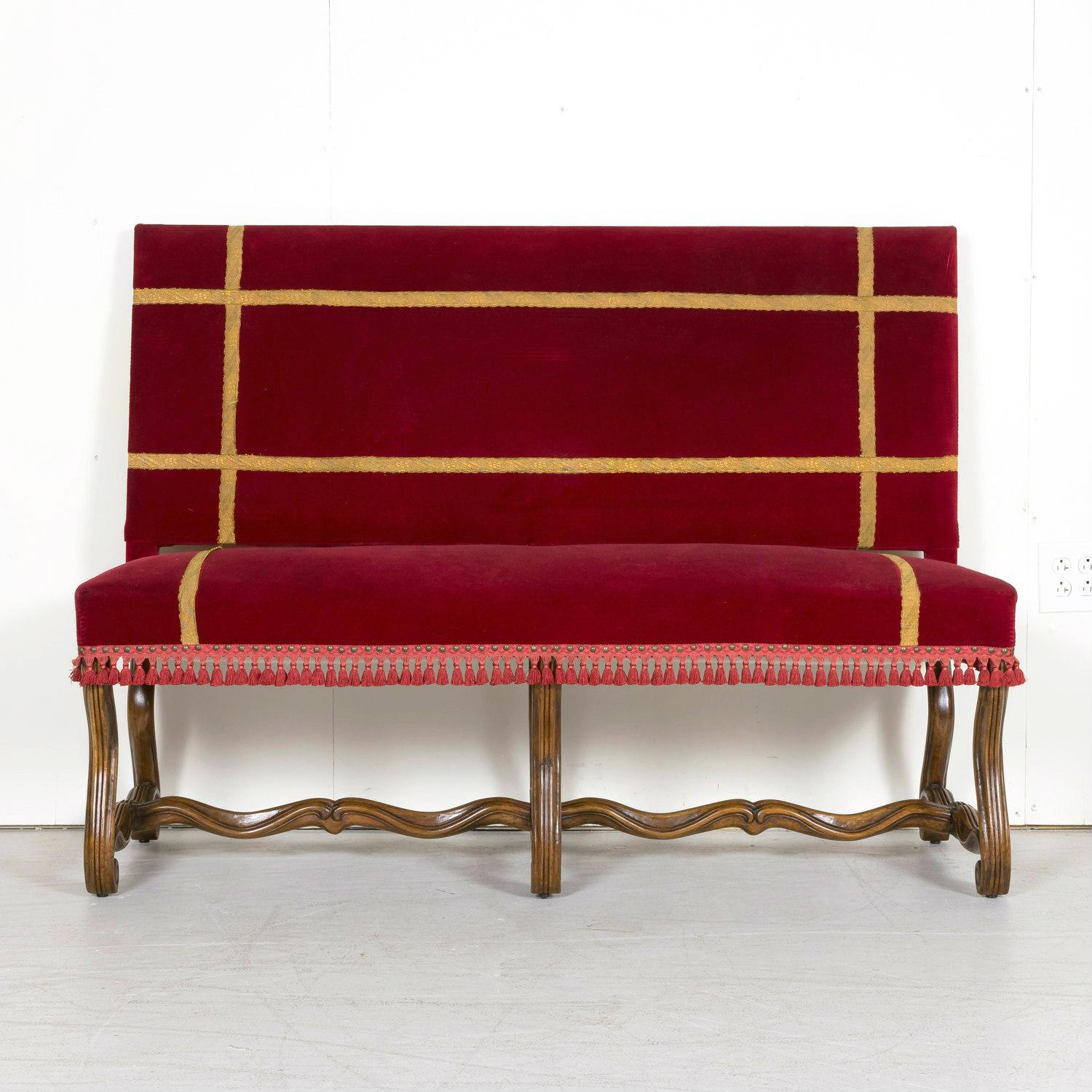 A 19th century Louis XIV style hand carved Spanish bench or banquette having original deep red velvet upholstery with metallic gold trim tape, brushed fringe, and nailhead trim, circa 1890s. This beautiful Spanish bench from the Catalan region is