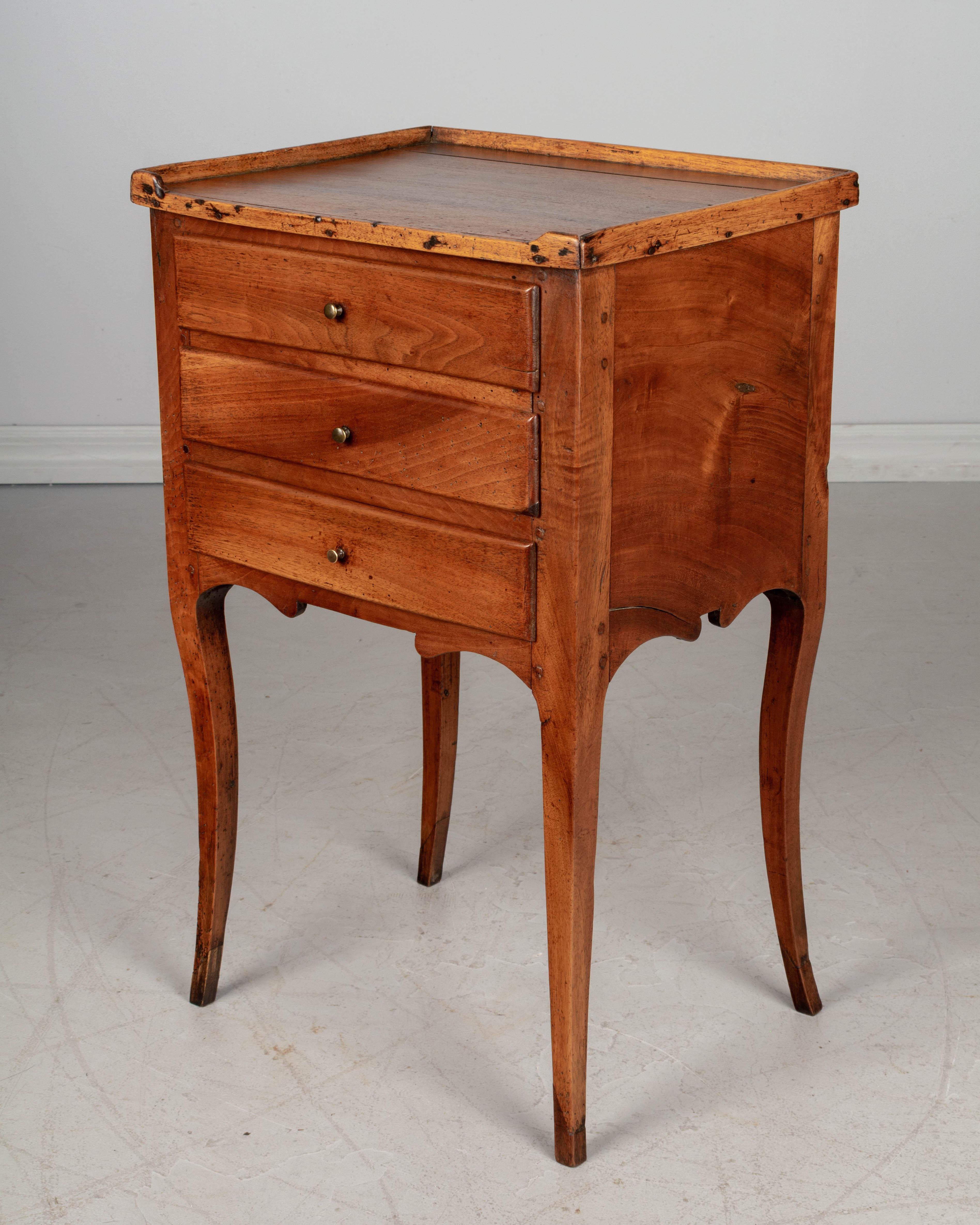 An early 19th century Louis XV style country French walnut side table, or nightstand, with curved legs and raised gallery. Three dovetailed drawers with small brass knobs. The interior of the top drawer is divided into small compartments.