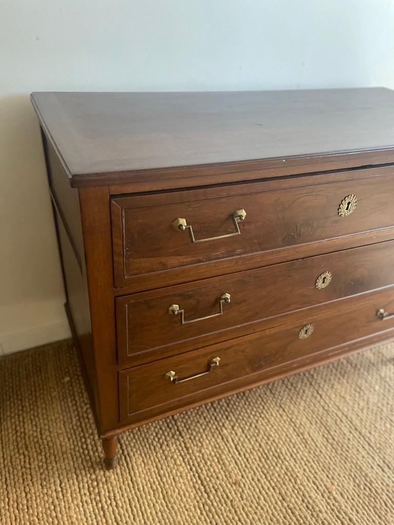 A French Louis XVI style walnut commode from the 19th century, with three graduated drawers, linear brass hardware, fluted side posts and cylindrical tapered legs. Created in France during the 19th century, this commode showcases the stylistic