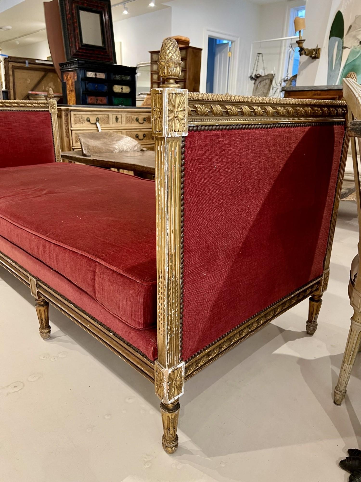 Este diván dorado Luis XVI del siglo XIX tiene una elegante silueta neoclásica, con detalles finamente tallados, patas estriadas y rosetones decorativos. La estructura de madera dorada muestra su pátina original, mientras que el asiento y los