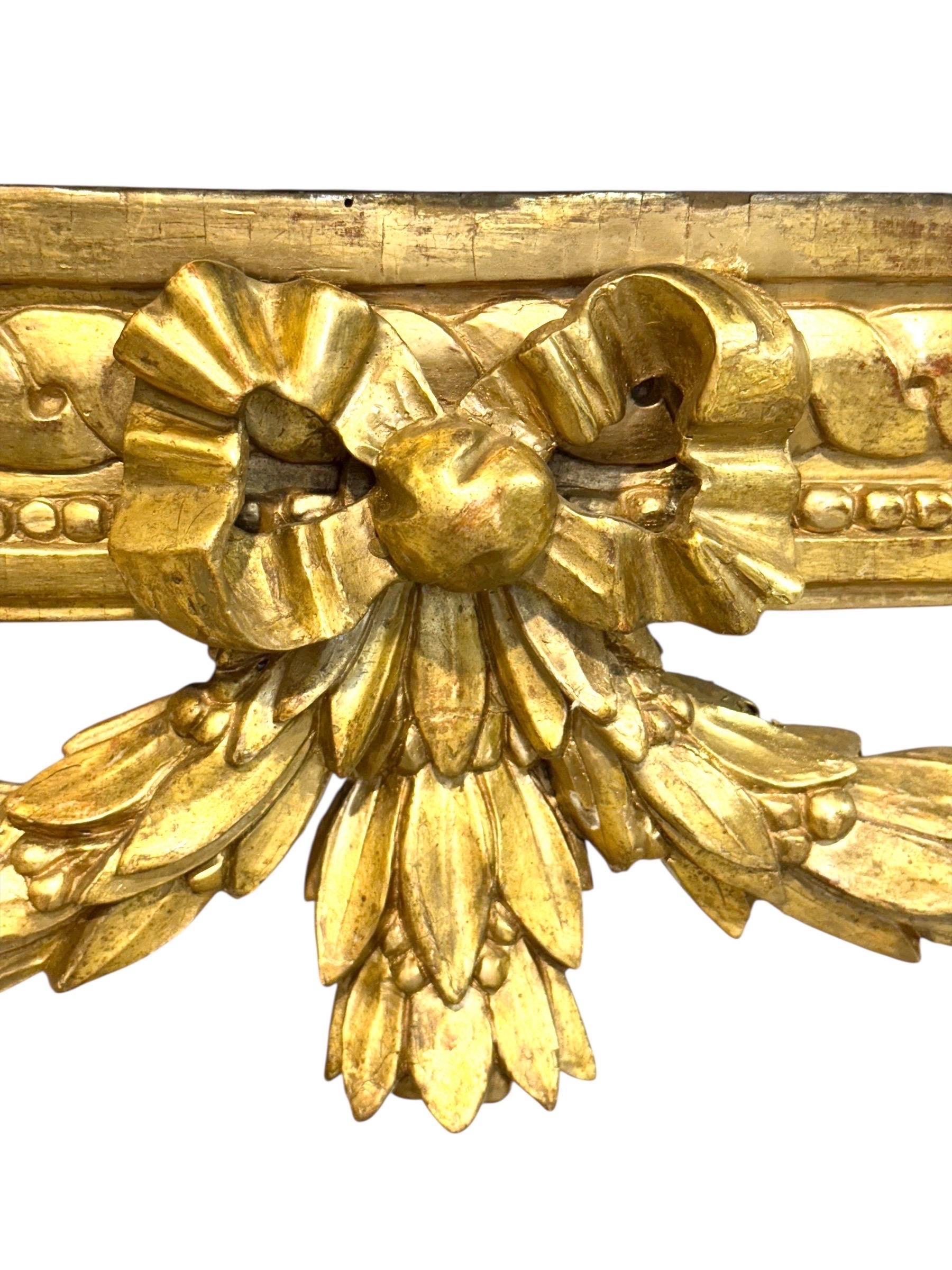 Fina consola de madera dorada Luis XVI de principios del siglo XIX, de forma demi-lune, nítidamente tallada y ricamente dorada. El friso perfilado está adornado con guirnaldas de hojas de laurel atadas por un lazo central, separadas por rosetas y