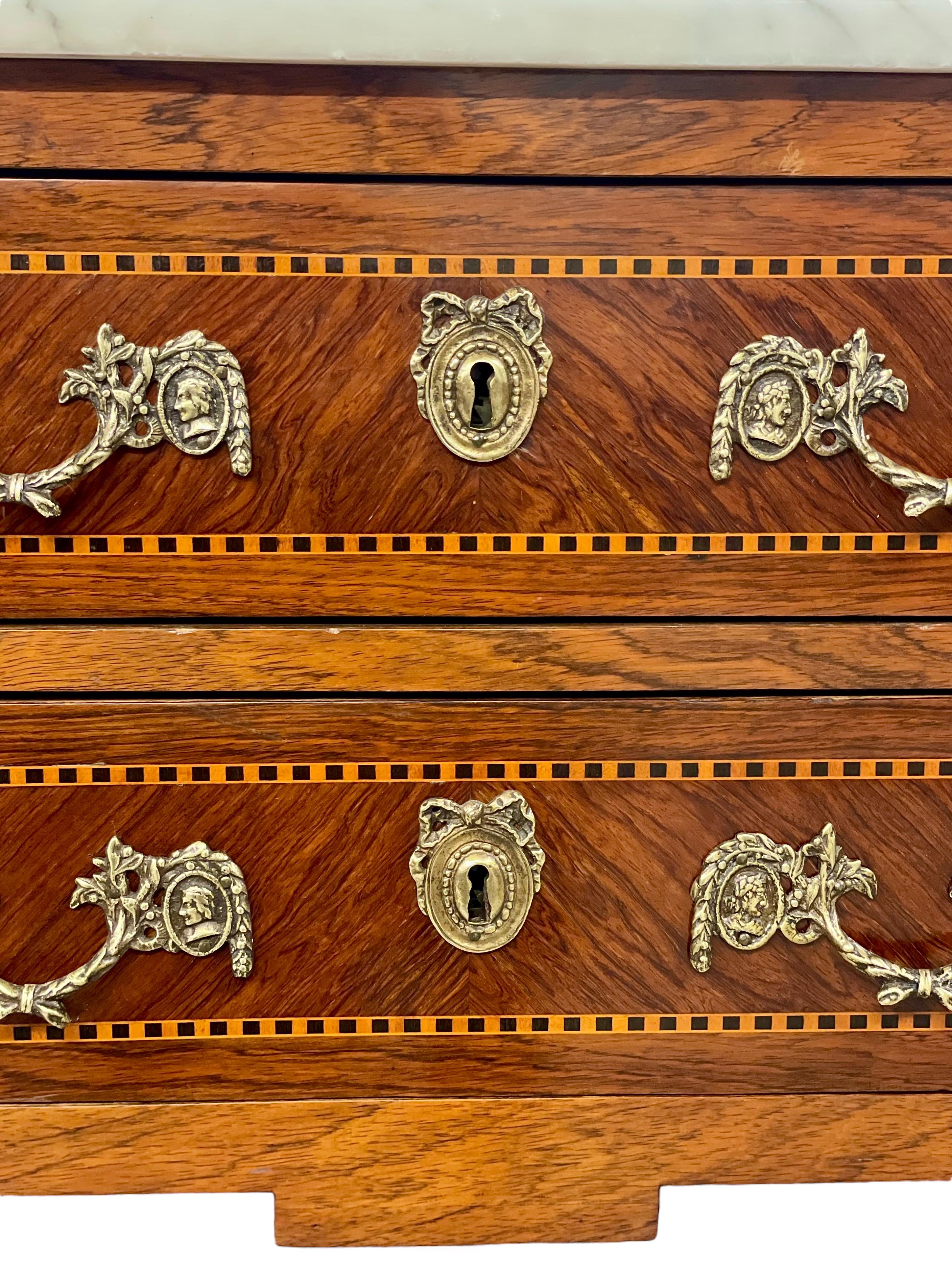 Eine zierliche und sehr elegante Kommode mit zwei Schubladen im Louis XVI-Stil aus furniertem Holz und Intarsien, mit Marmorplatte und verzierten vergoldeten Messingbeschlägen. Bei dieser Kommode mit dem kuriosen Namen stehen immer zwei Schubladen