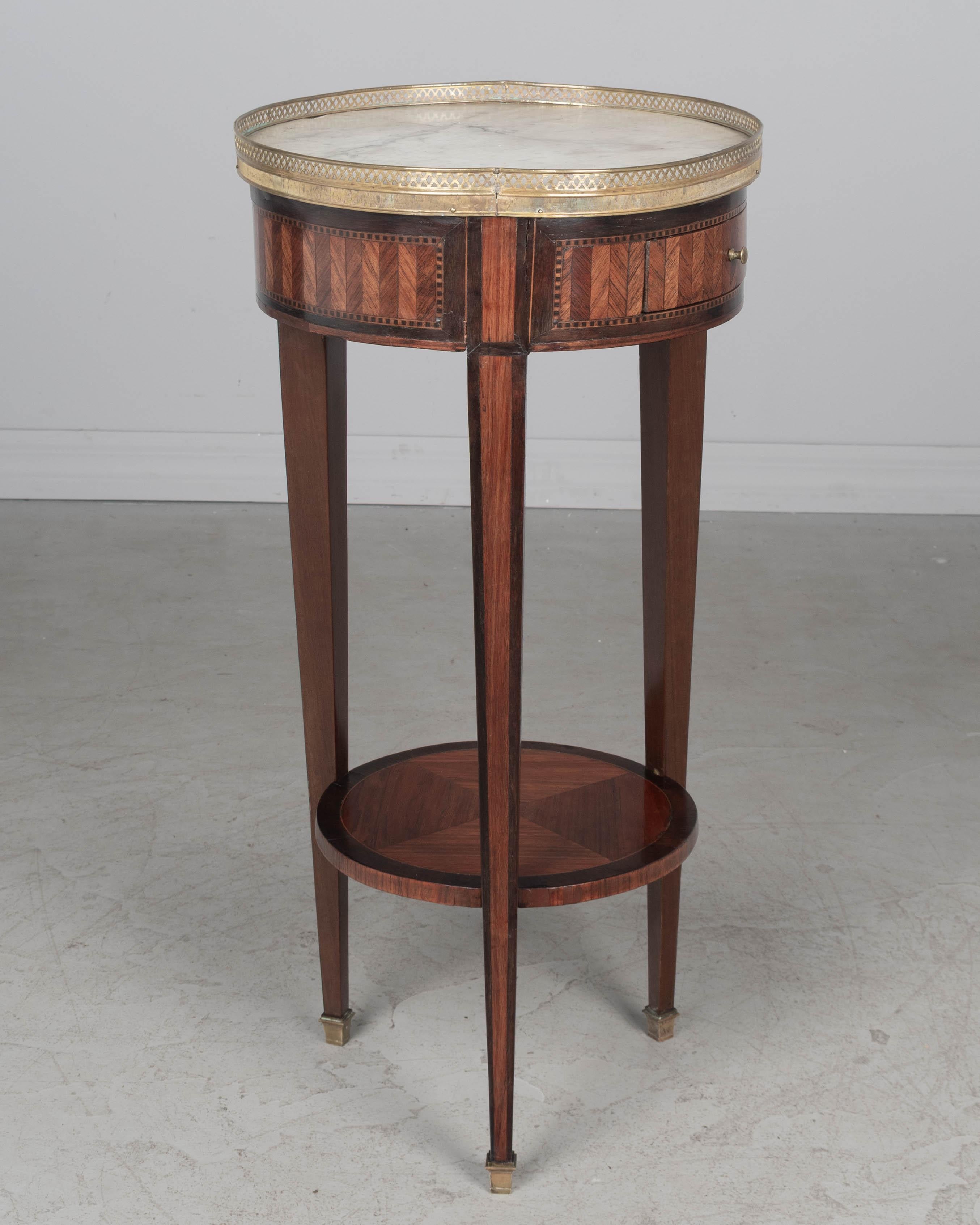 A mid 19th century French Louis XVI style marble top marquetry circular side table with a dovetailed drawer and lower shelf. Made of veneer of mahogany and various exotic wood veneers. White marble top with bronze gallery. Slender tapered legs with