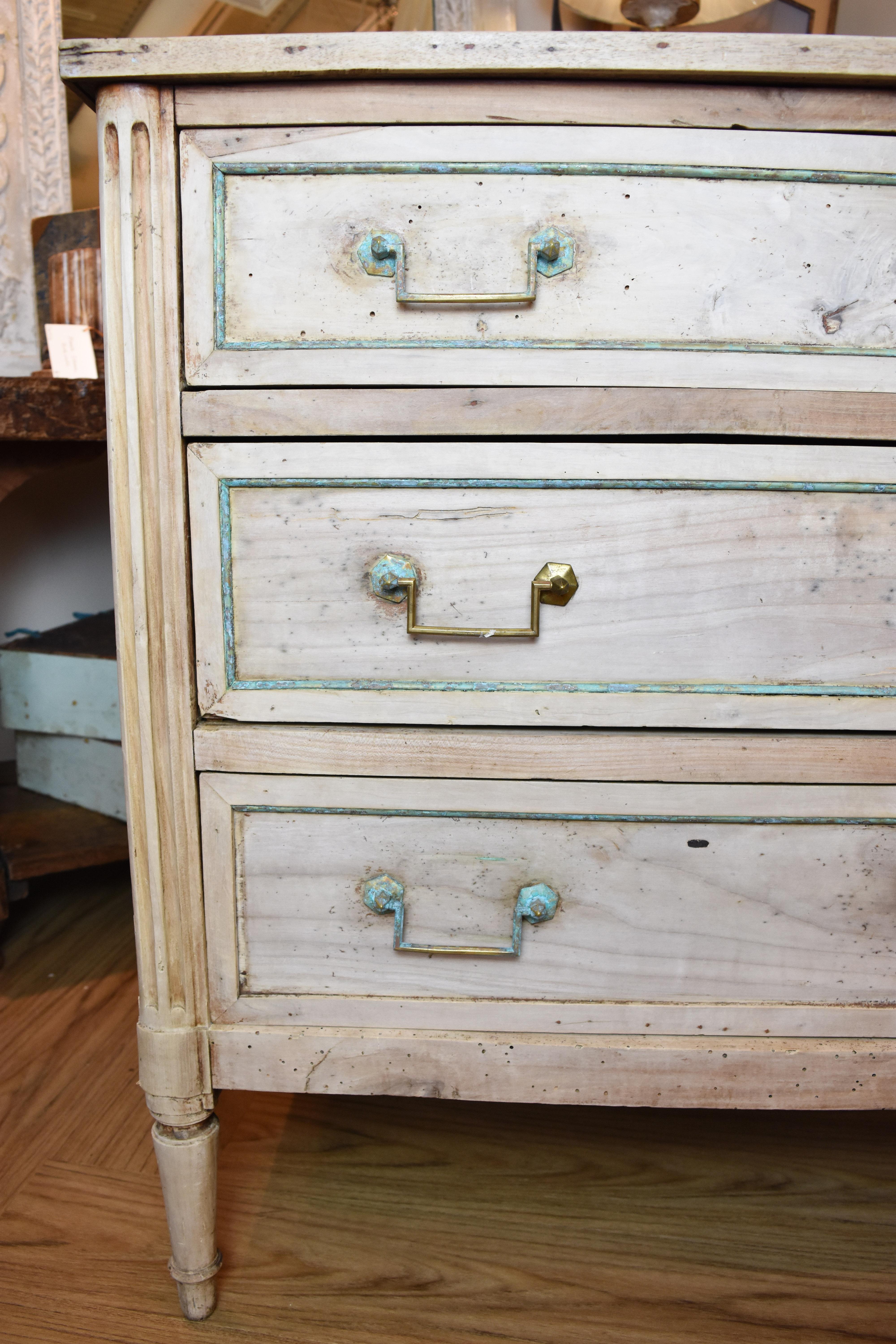 This 19th century bleached commode has three functional drawers with drop pulls. The bronze pulls and brass trim surrounding the drawers and side panels has oxidized and is a lovely blue/green color. The bottom drawer is missing some of the bronze