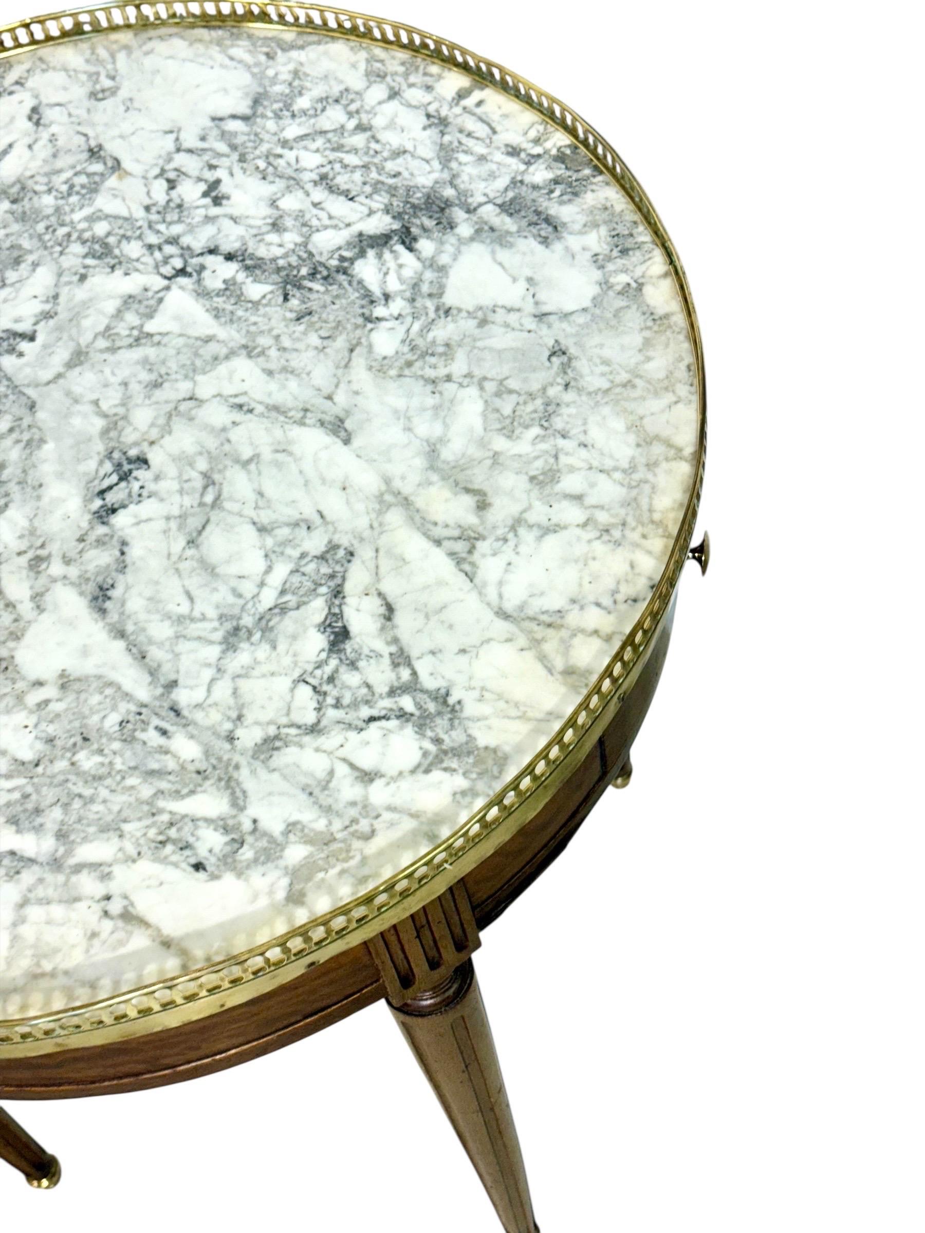 Cette table bouillotte de style Louis XVI du XIXe siècle illustre le raffinement du design néoclassique français. Fabriqué en noyer à la patine chaude et élégante, il présente un plateau rond en marbre blanc joliment veiné de gris, entouré d'une