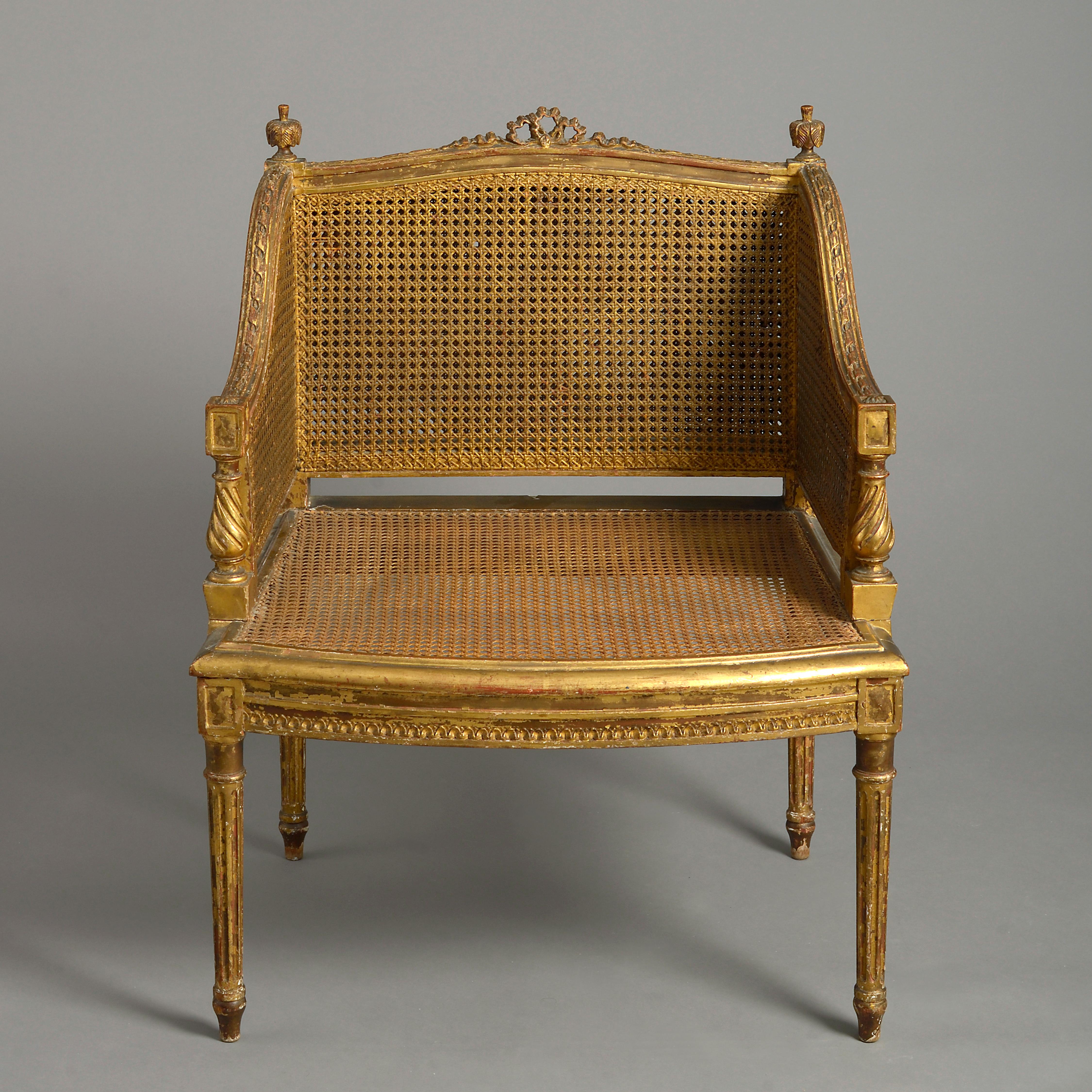 A late 19th century carved giltwood bergère in the Louis XVI manner, the frame with ribbon carved back rail and finials, having canework throughout and raised on turned tapering legs.