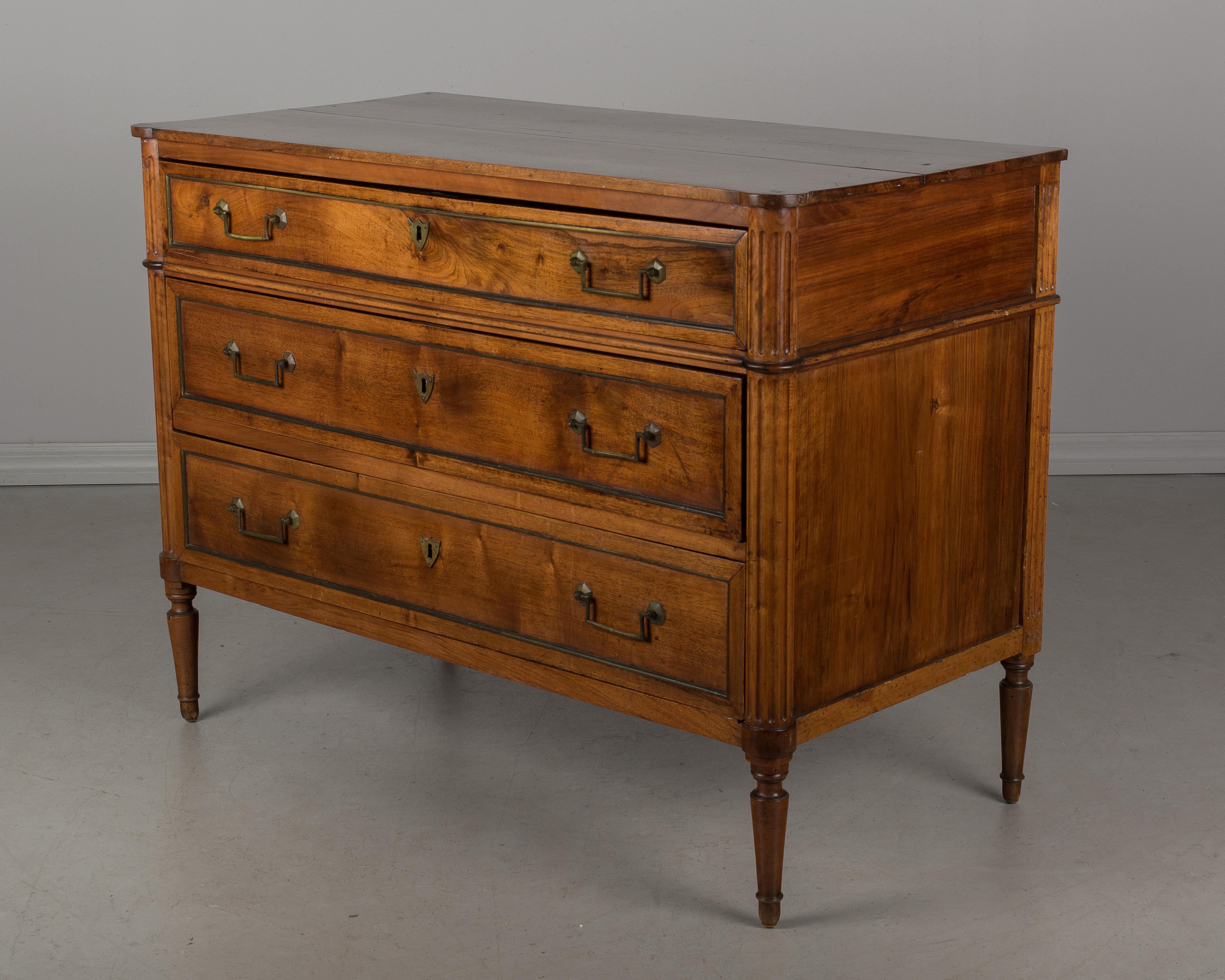 A 19th century French Louis XVI style commode made of solid walnut. Three dovetailed drawers with original brass trim, pulls and escutcheons. There are no locks or keys. Fluted corners with shaped top. Pegged construction. Top drawer is slightly