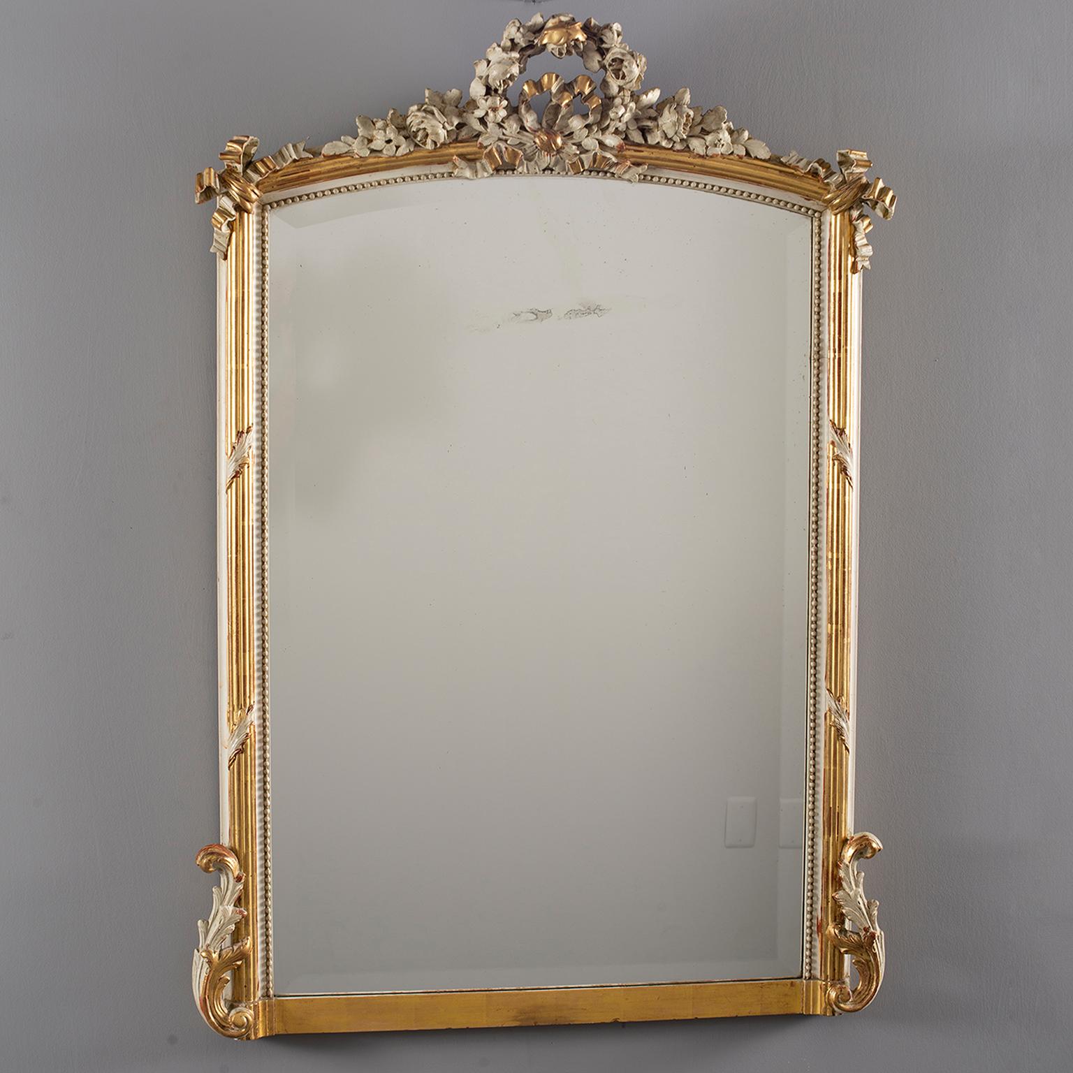French Louis XVI style mirror has wood frame with cream color and gilded finish. Detailed decorations at bottom corners and wreath form crest, circa 1880s. Beaded inner edge, mirror is newer. Overall very good antique condition with some scattered