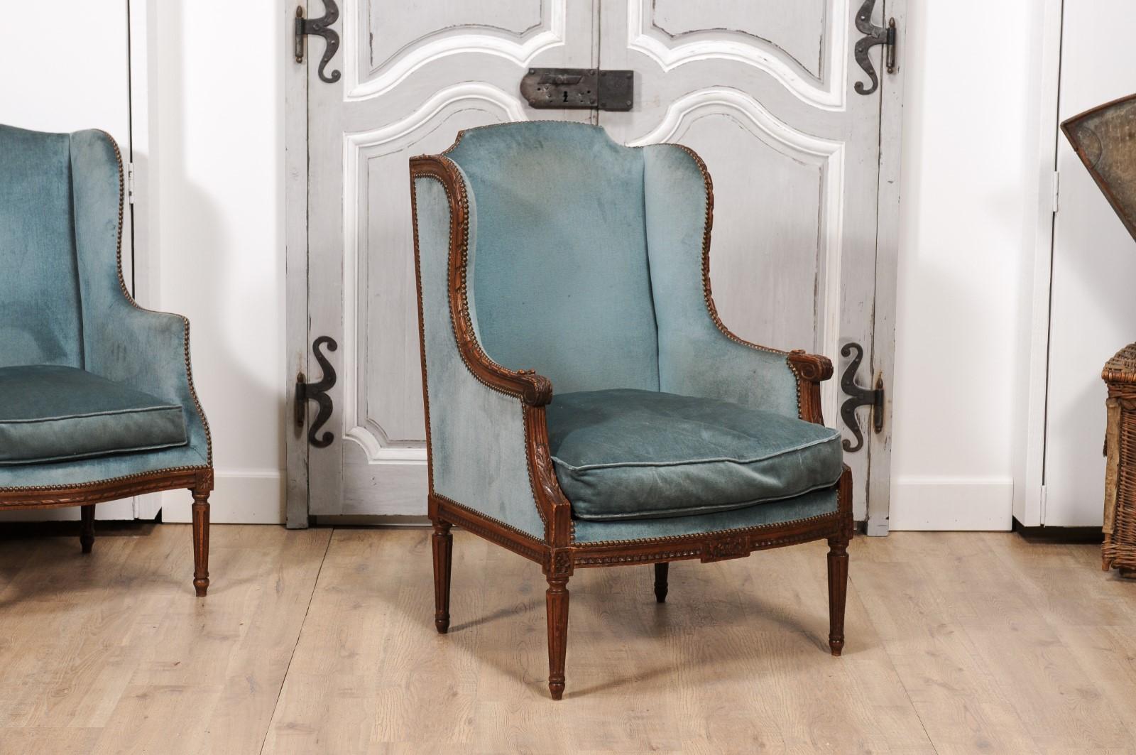 A large French Louis XVI style walnut wingback bergère chair from the 19th century with carved foliage décor and old blue velvet upholstery. Introducing a French Louis XVI style bergère chair, dated to the 19th century, imbued with a rich historical