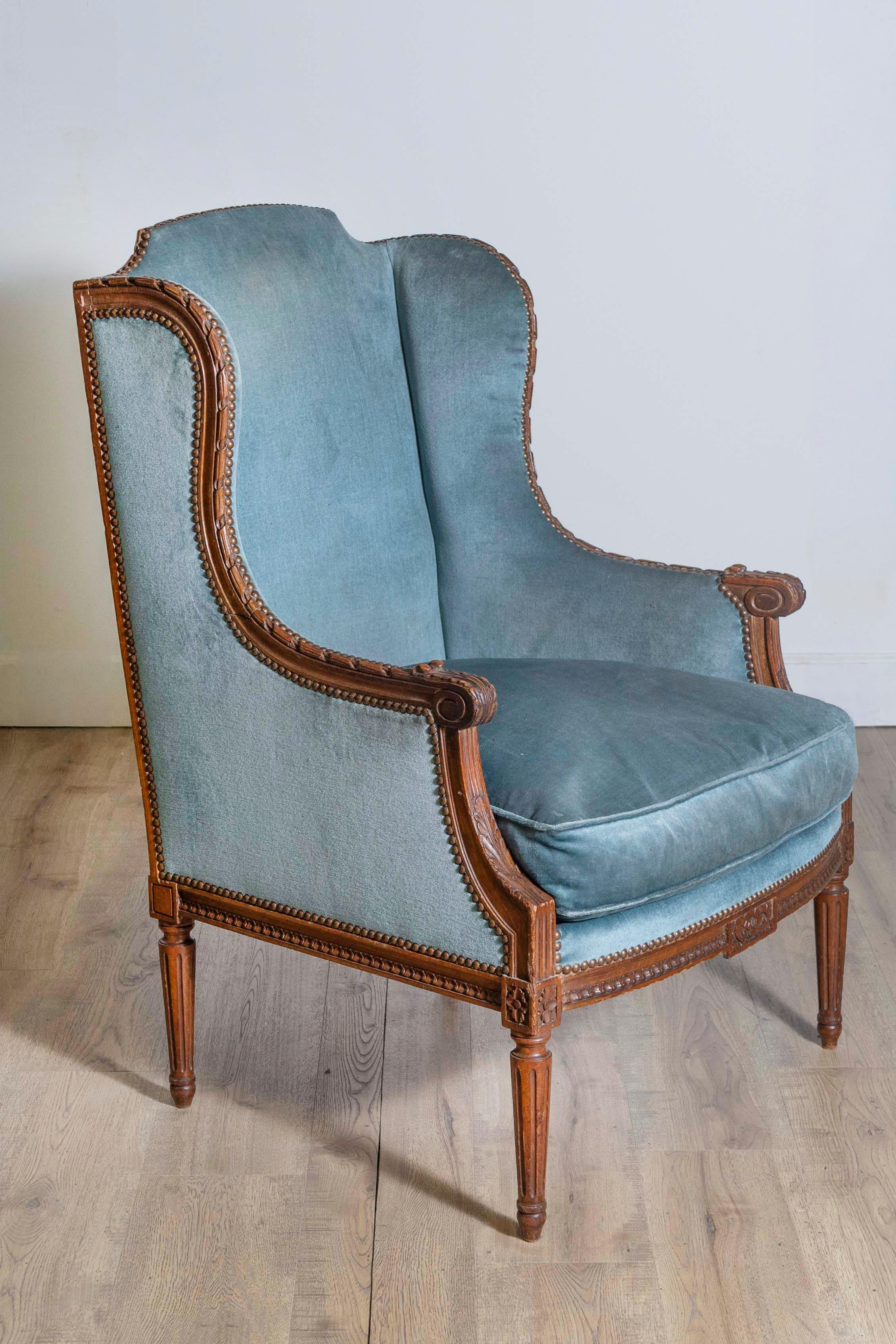 A large French Louis XVI style walnut wingback bergère chair from the 19th century with carved foliage décor and old blue velvet upholstery. Introducing a French Louis XVI style bergère chair, dated to the 19th century, imbued with a rich historical