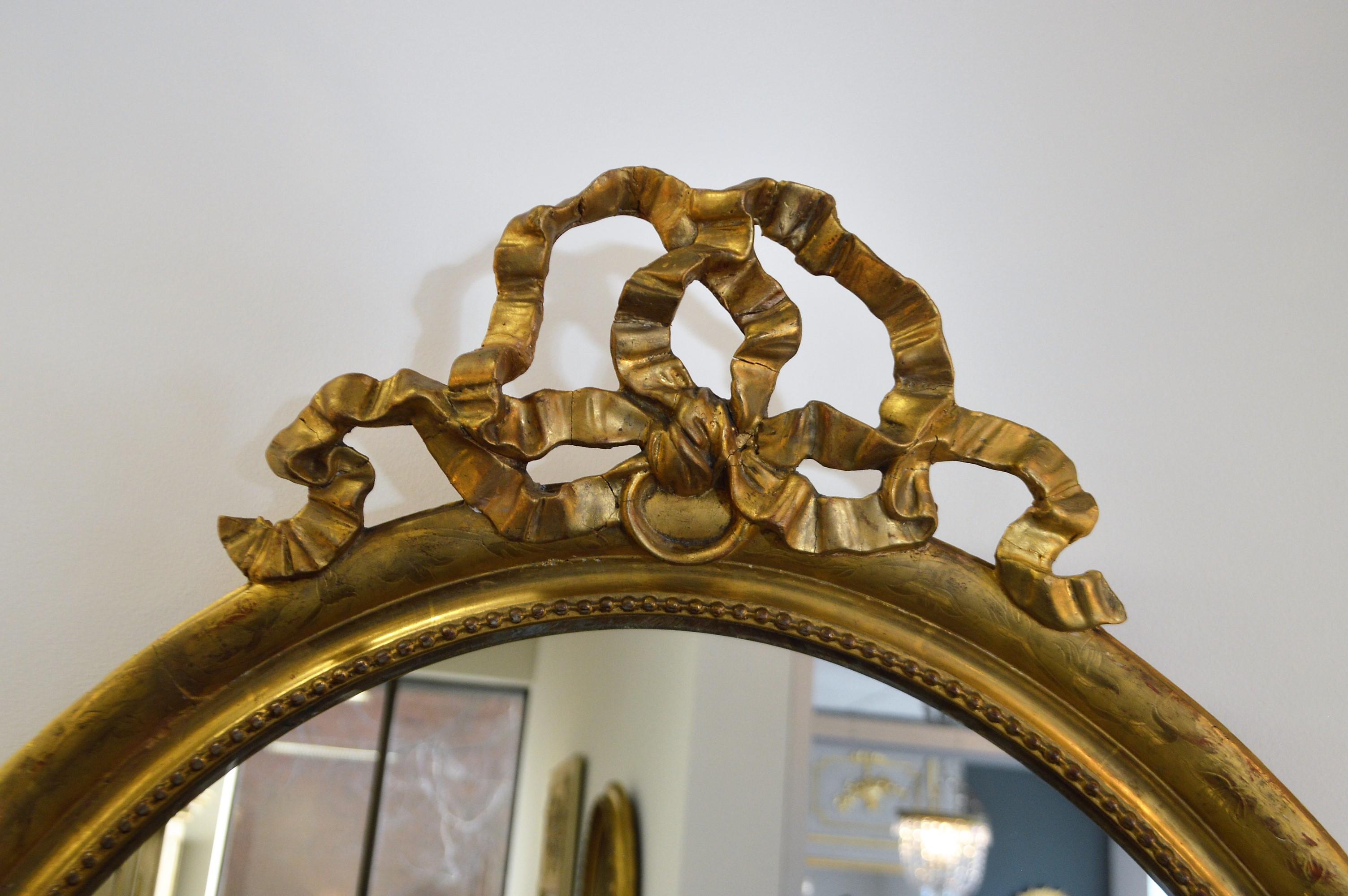 Louis XVI style gilded oval mirror from 19th century France, large bow at top of the mirror, with delicate floral design and pearling all around the surface of the frame.