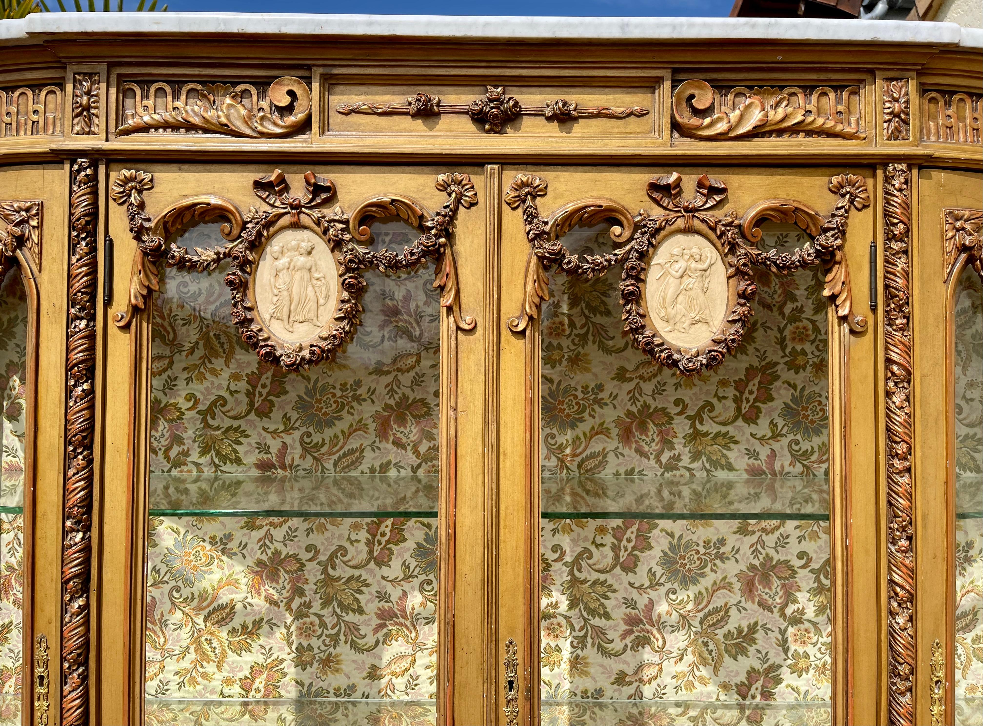 Magnifica vetrina curva in legno dorato con piano in marmo bianco in stile Luigi XVI. Questa vetrina si apre con due porte anteriori ornate da ghirlande di fiori e medaglioni Wedgwood con scene antiche come decorazione, e si apre anche su entrambi i